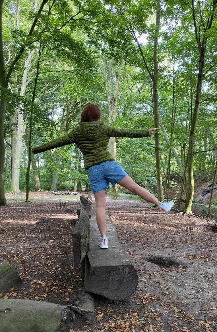 Person balancing on a wooden log in a forest, with arms extended to the sides, wearing a green jacket, blue shorts, and white sneakers.