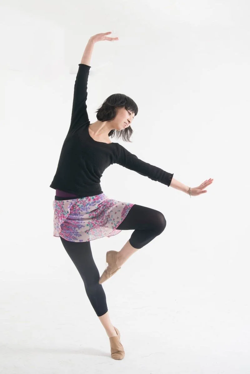 A woman dancing or performing a ballet pose on a white background, wearing a black top, a colorful floral skirt, black leggings, and beige dance shoes.