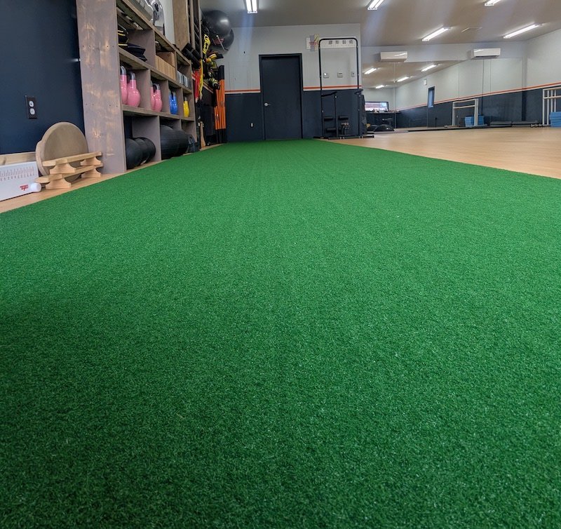Interior of a gym with a green turf and workout equipment along the wall, bright fluorescent lights, and a mirrored wall.