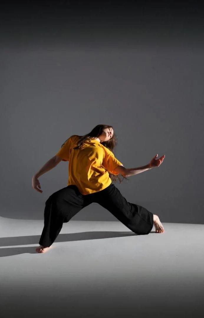 A woman in a yellow shirt and black pants performing a dance pose on a gray background.