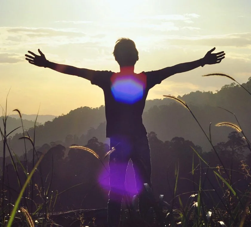 Person standing in a field with arms outstretched towards the sunset over hills and trees.
