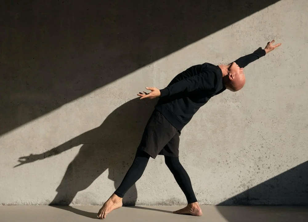 Person doing a yoga pose against a wall with shadows cast on the wall. Outdoor expressive movement training at La Muse Studio