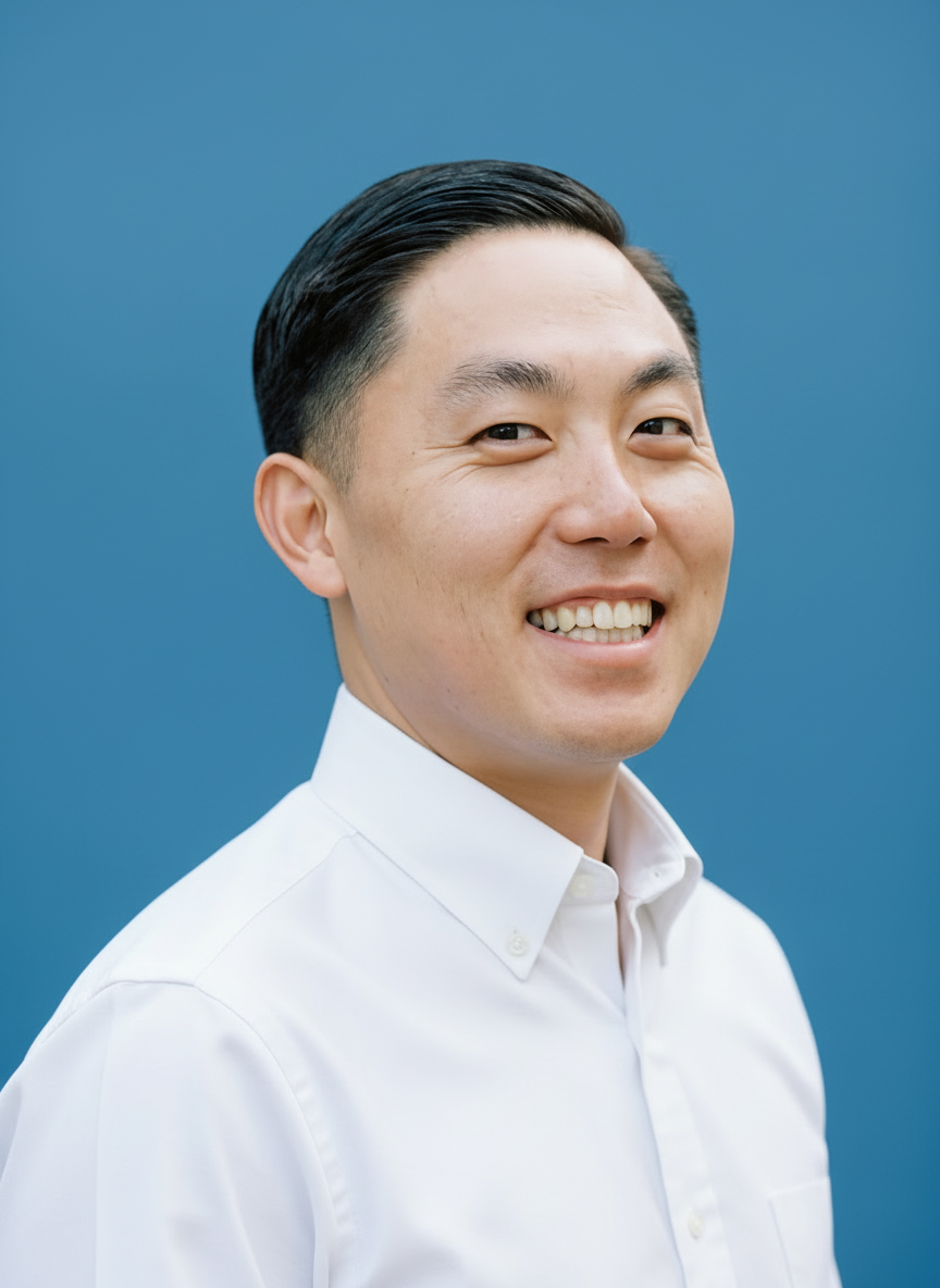A smiling Asian man in a tuxedo and bow tie standing outdoors with greenery in the background.