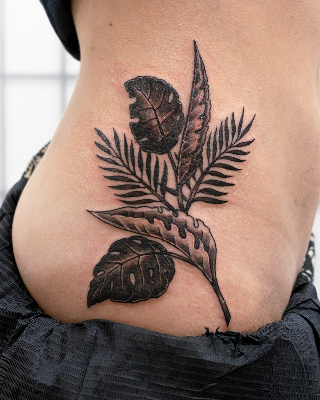 A tattoo of various leaves including ferns and other foliage in black and gray ink on a person's side torso.