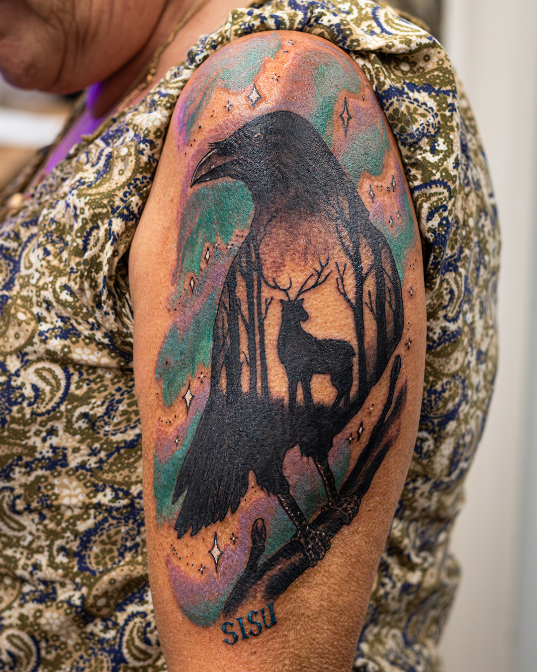 Tattoo of a black crow, with a forest scene including a deer and trees, on a person's upper arm surrounded by colorful clouds, stars, and sparkles.