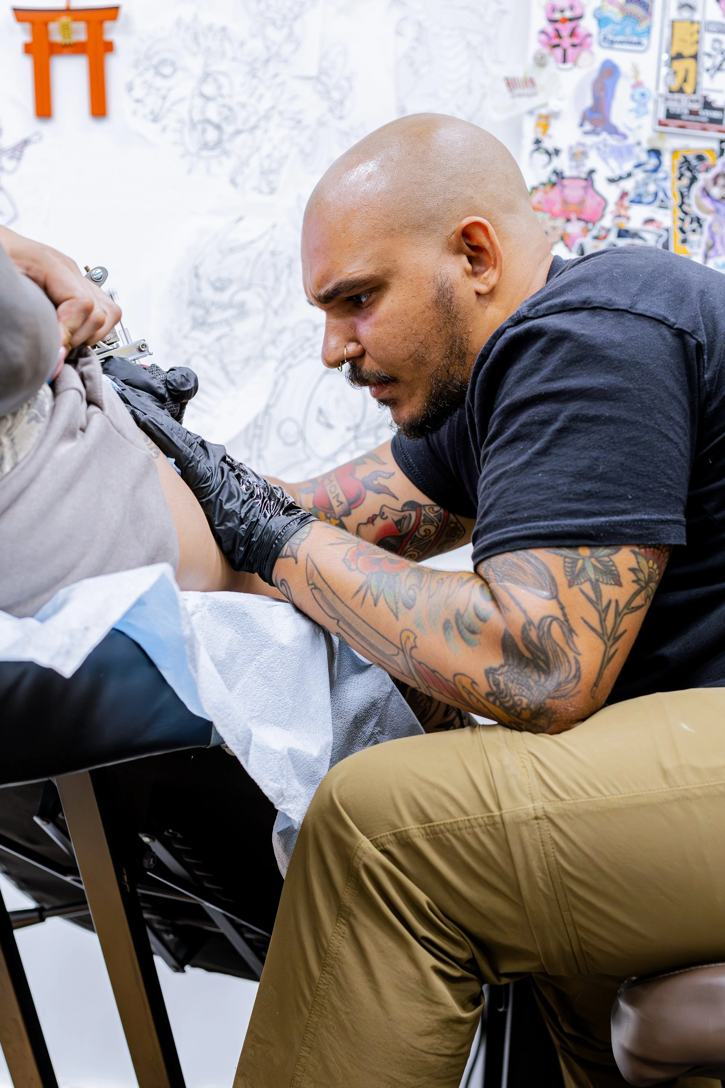 Tattoo artist in black shirt and beige pants tattoos a client's arm in tattoo studio with posters and sketches on the wall.