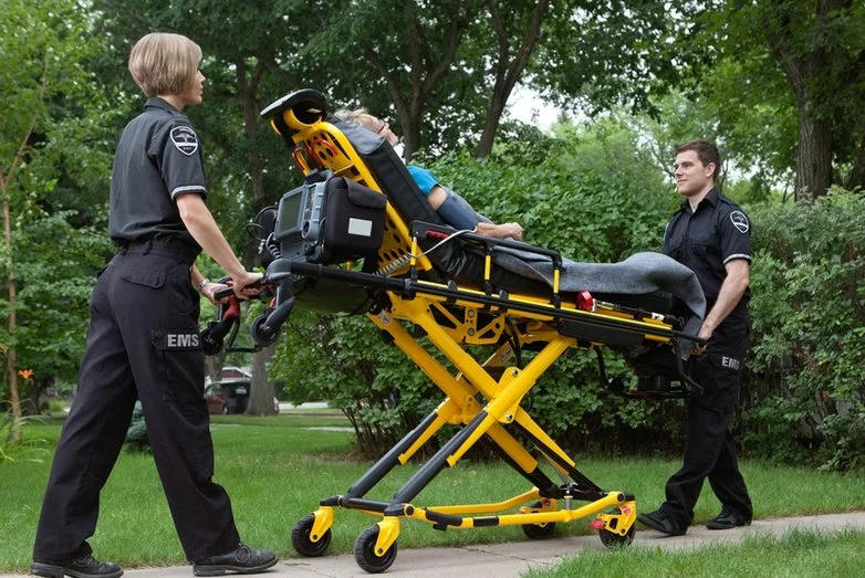 Two emergency medical technicians tow a patient on a stretcher through a park.