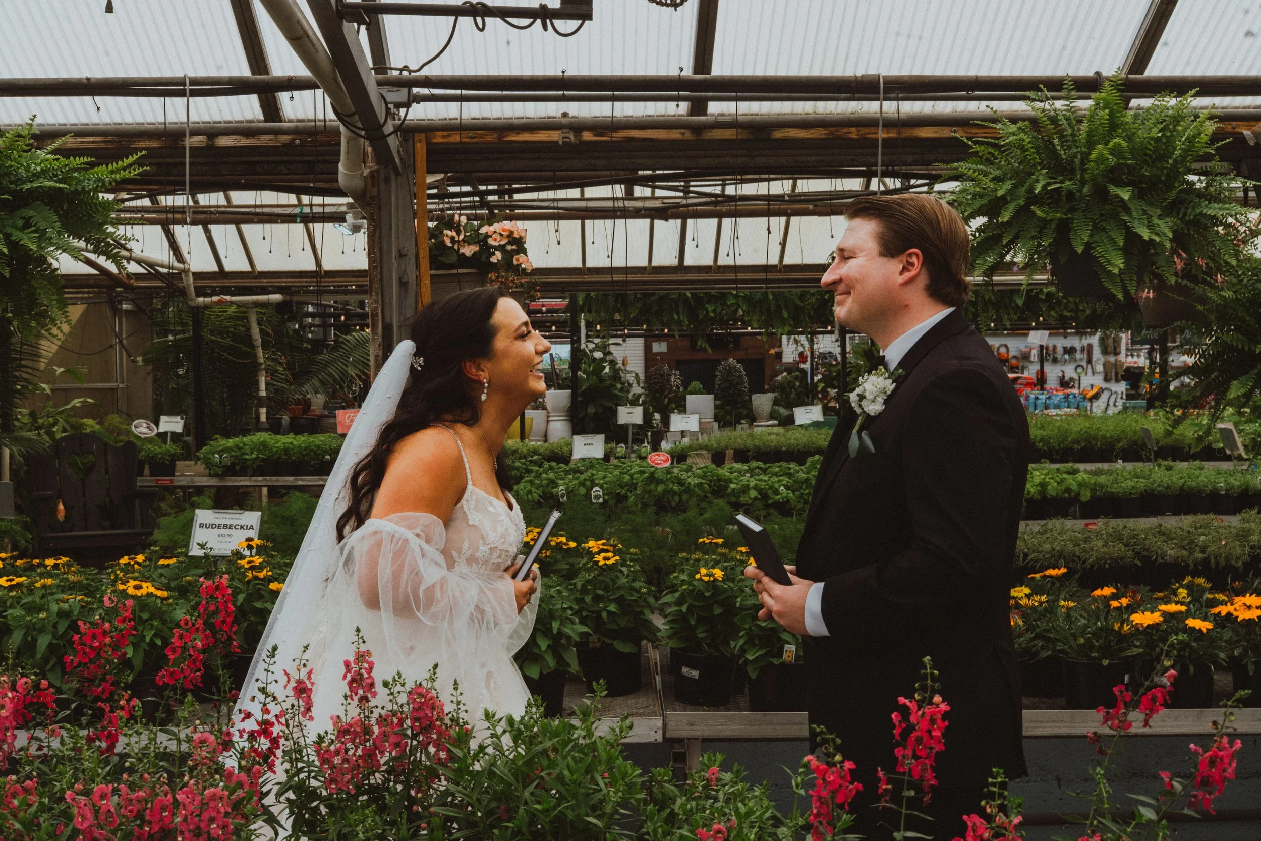 A bride and groom are smiling at each other in a greenhouse or garden center, surrounded by colorful flowers and plants during their wedding.