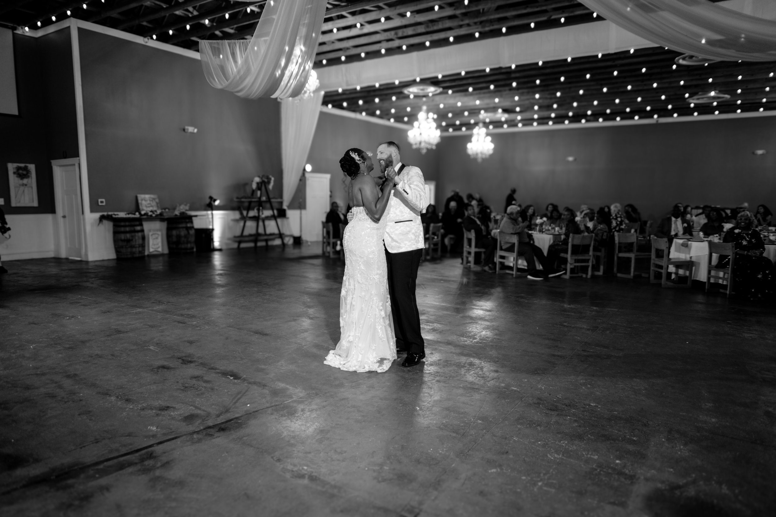 Black and white photo of a bride and groom dancing at their wedding reception in a decorated hall with string lights and chandeliers.