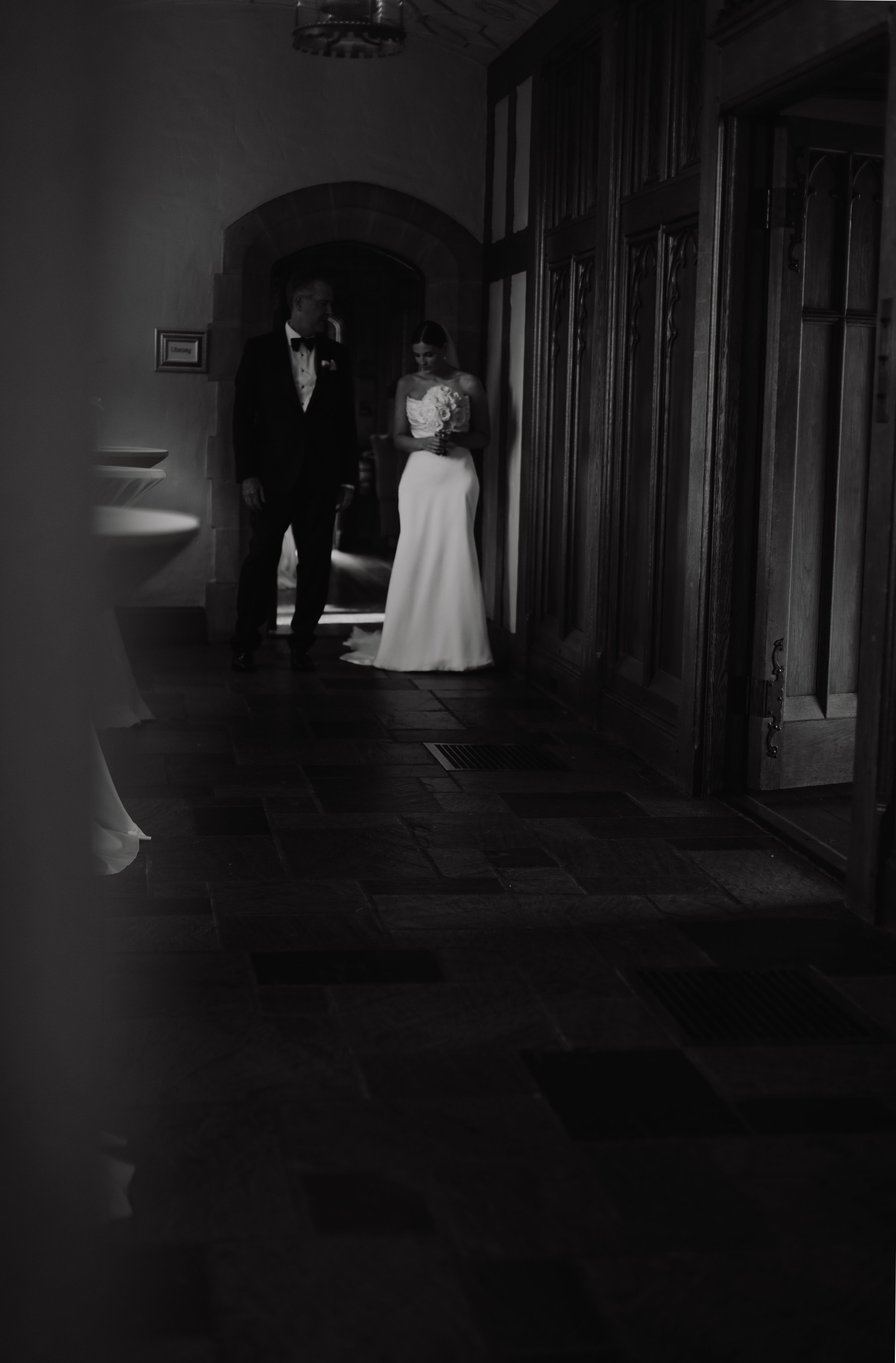A bride in a wedding dress holding a bouquet standing next to a groom in a tuxedo inside a dimly lit room.