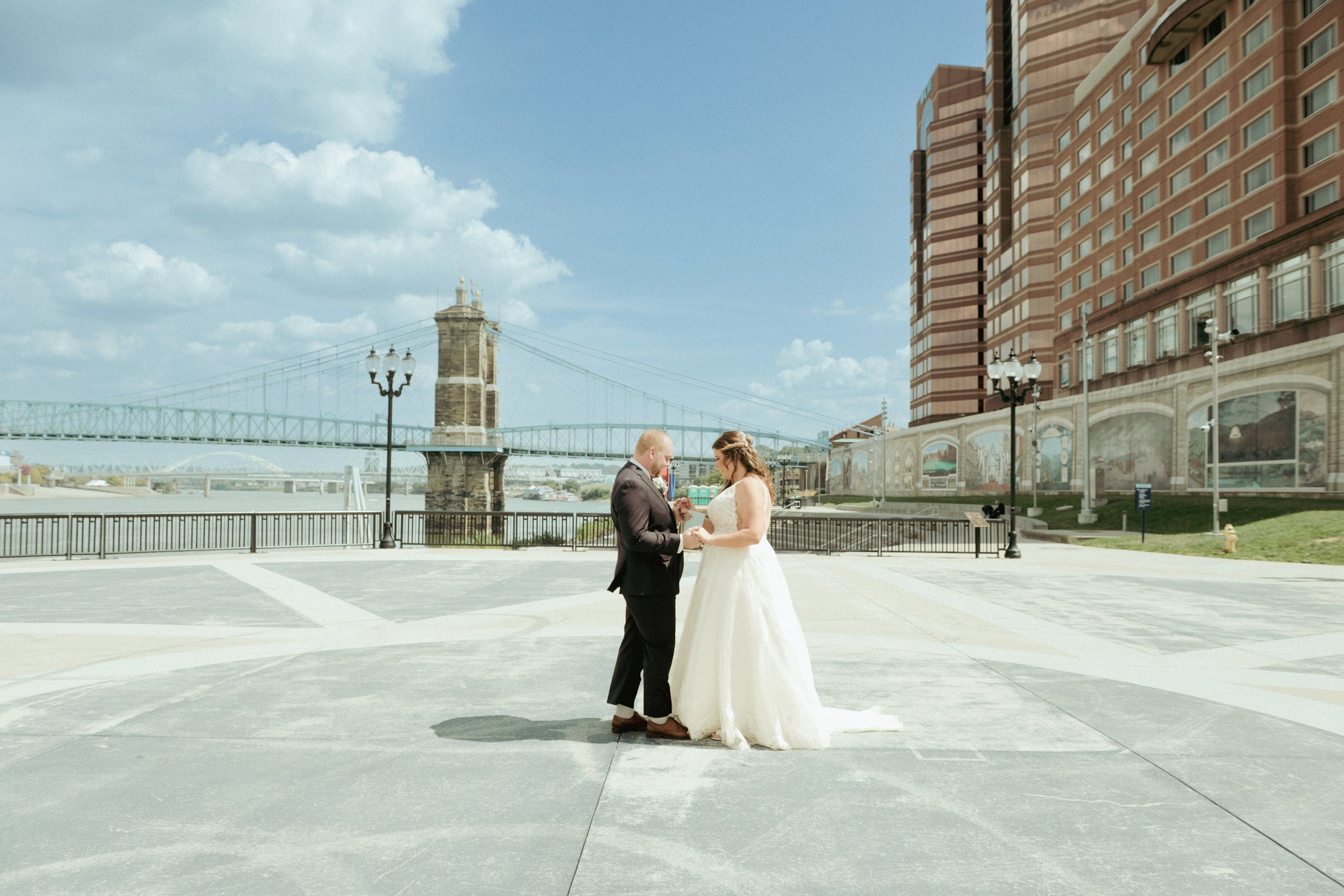 A bride and groom exchanging rings outside near a river, with a bridge and tall buildings in the background on a sunny day.
