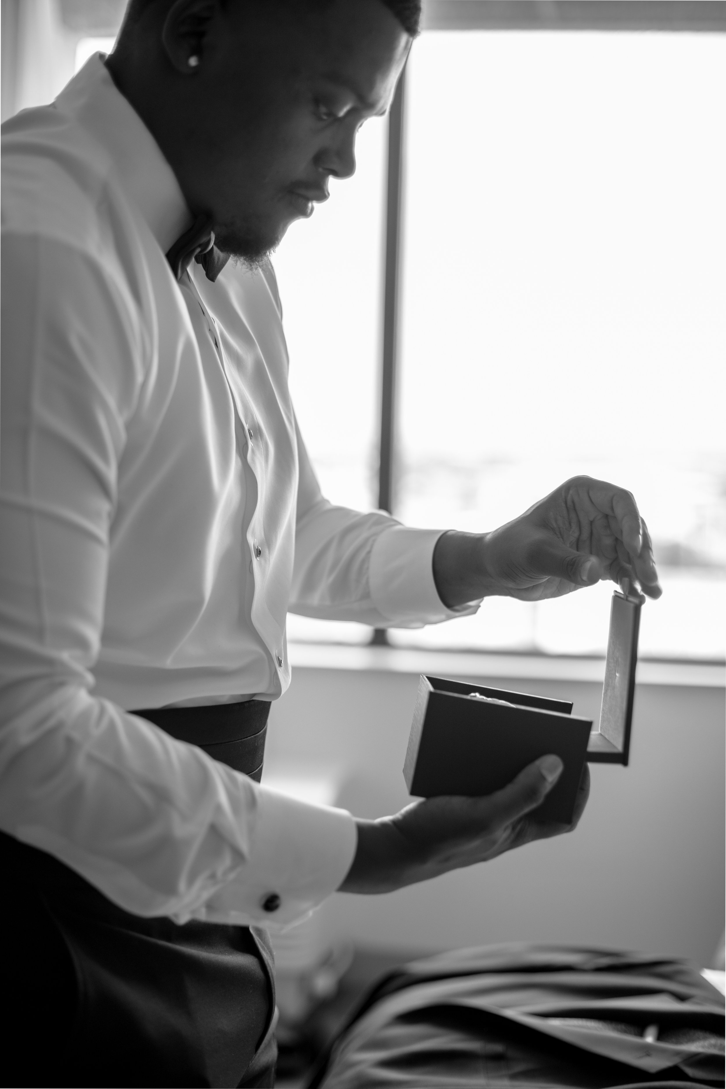 A man in a suit opening a small box, possibly a jewelry box, near a window.
