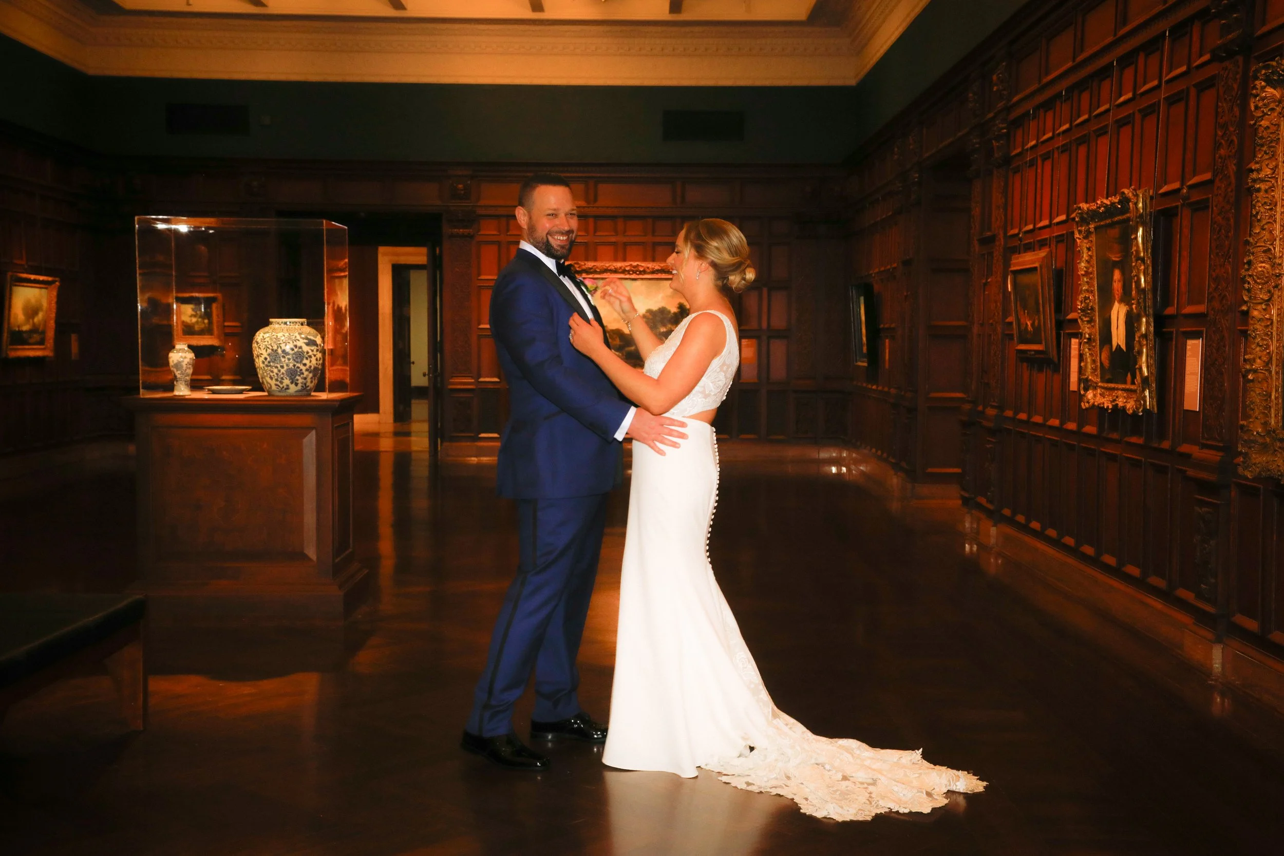 A bride and groom dancing in a wood-paneled room with artwork and vases on display.