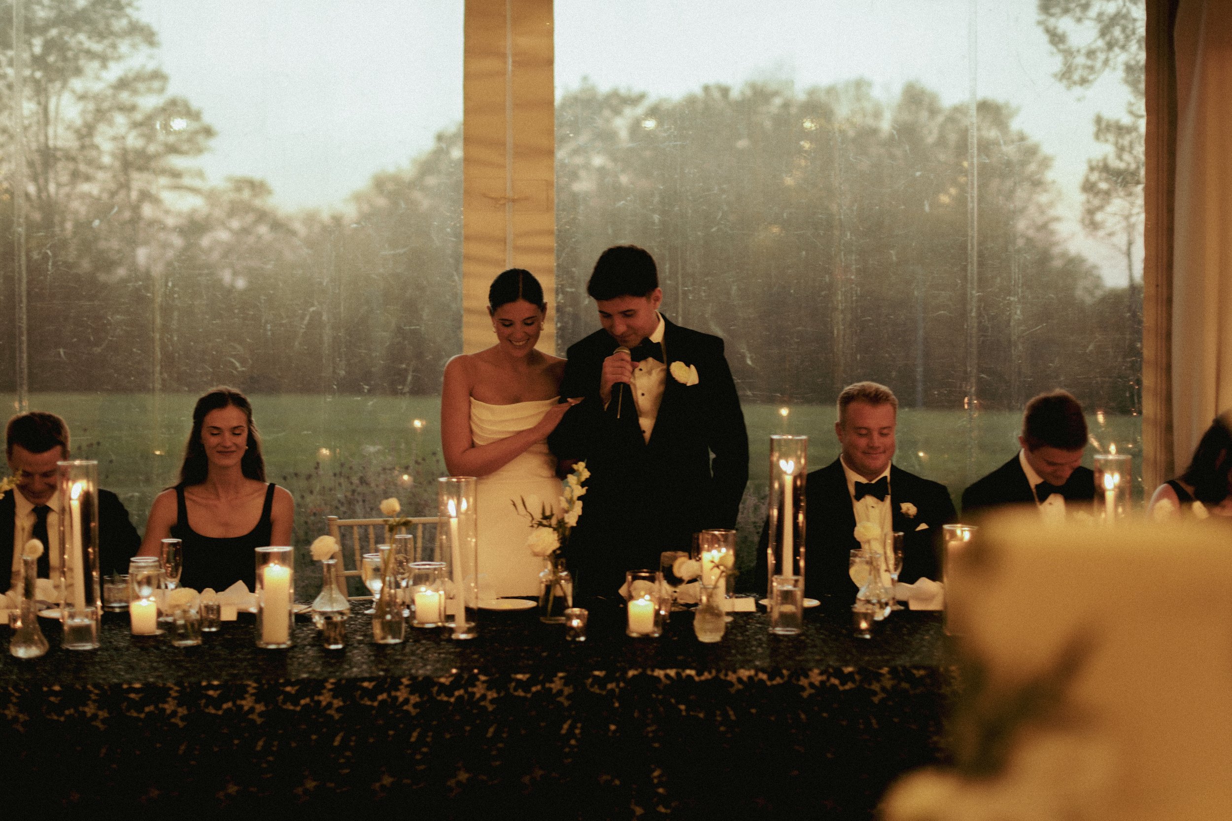 A wedding reception scene with a bride and groom standing and speaking into a microphone, surrounded by seated guests, with candles and floral centerpieces on the table.