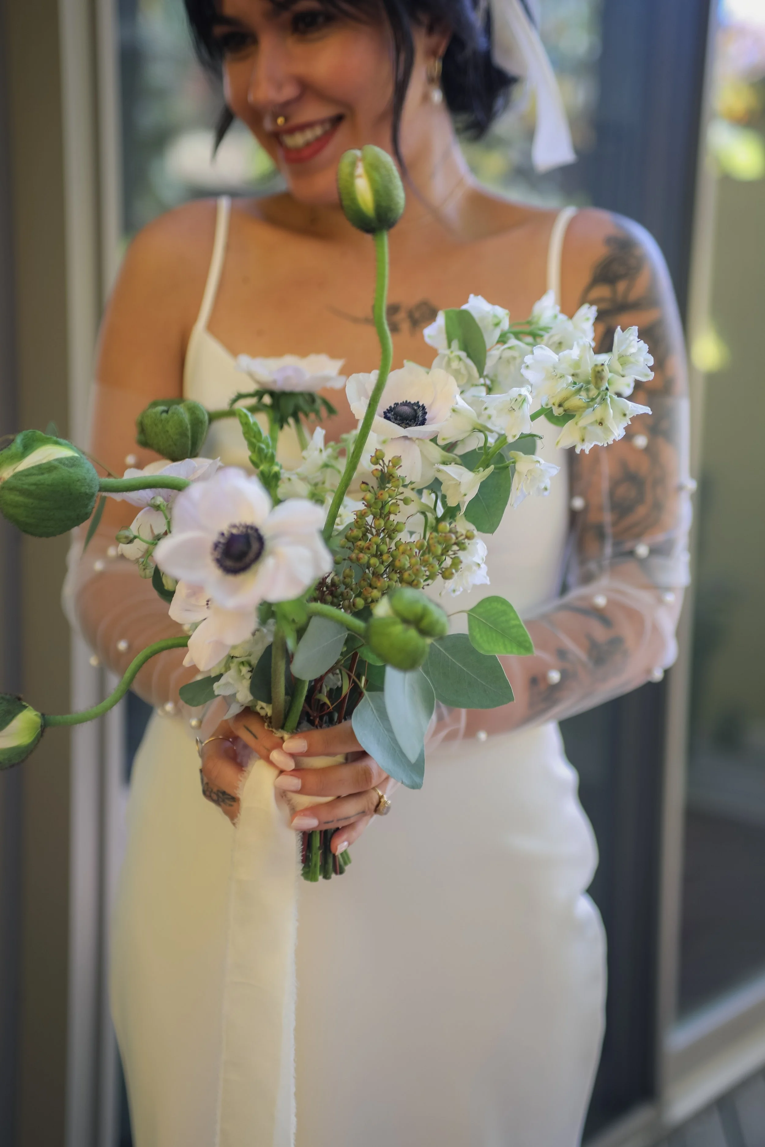 A woman in a white dress holding a bouquet of white flowers with green foliage, standing indoors near a window, smiling.