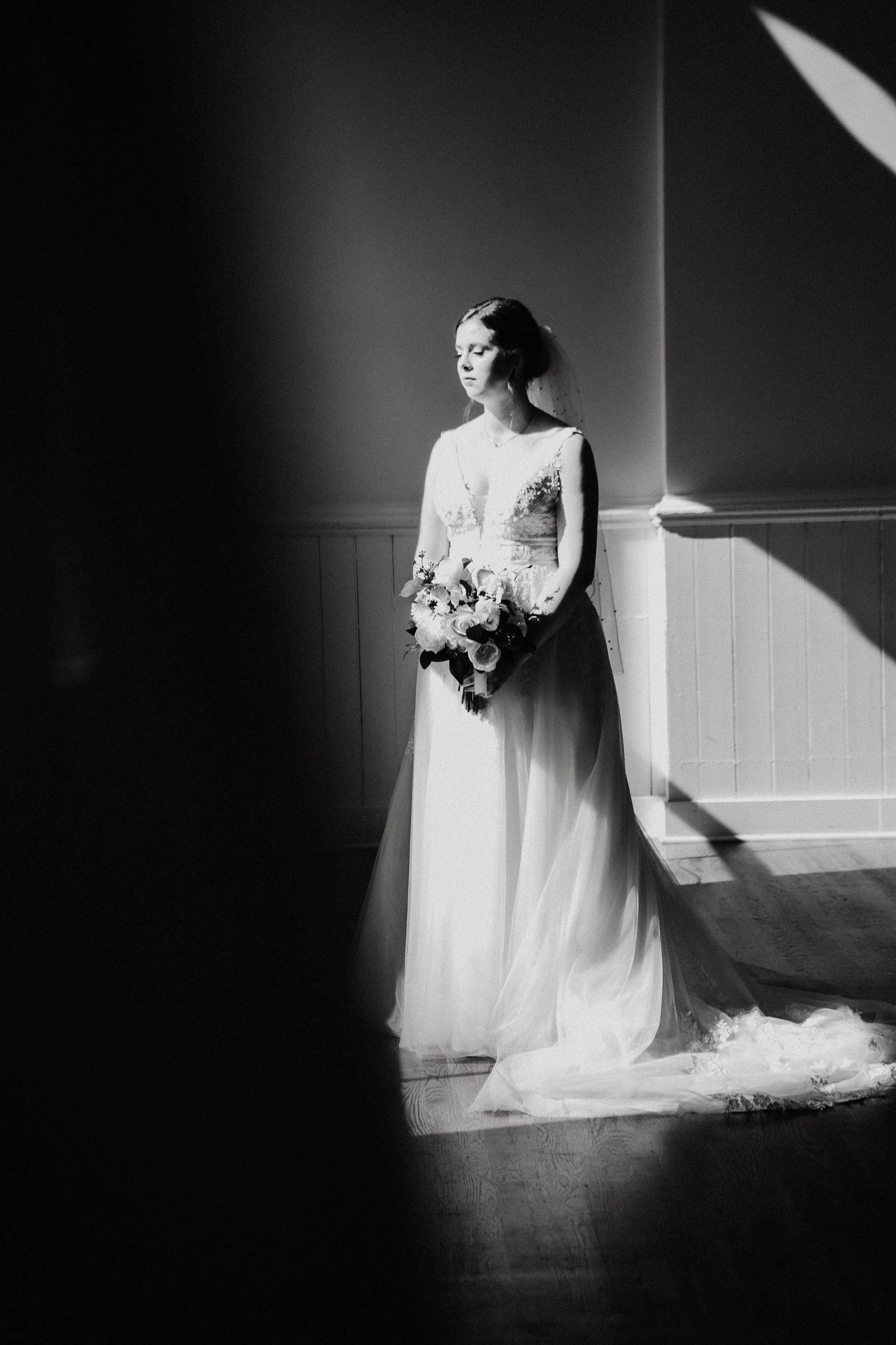 Black and white photo of a bride holding a bouquet, standing with eyes closed in a spotlight, with a plain wall background.
