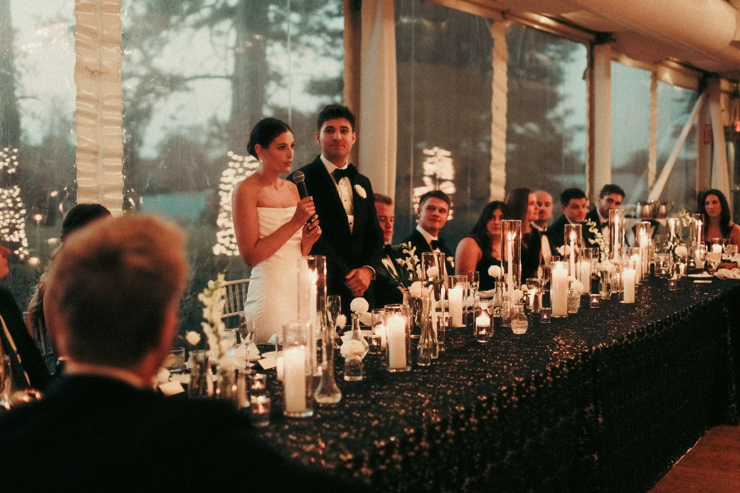 A bride in a white strapless wedding gown holding a microphone, standing beside the groom in a black tuxedo at a wedding reception. They are at a long decorated table with candles and flowers, with several guests seated behind them, in an elegant ven