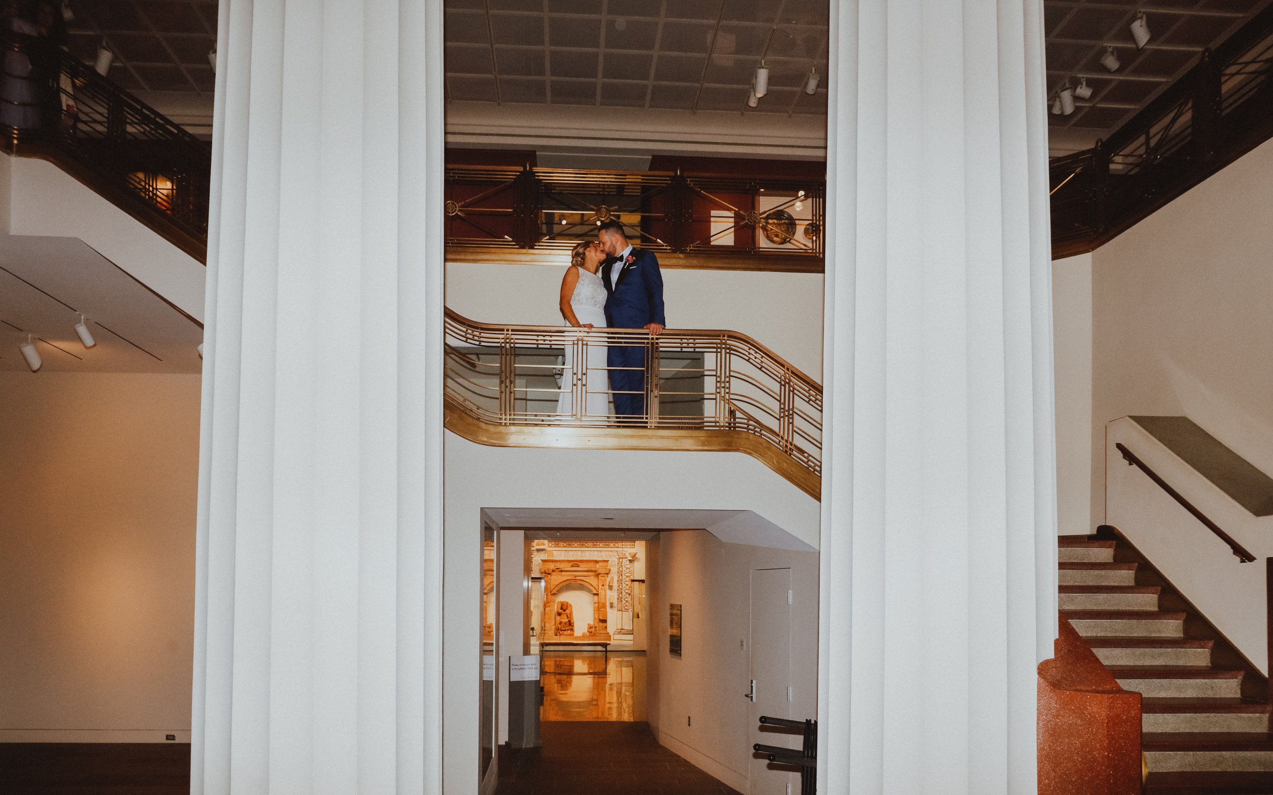 A bride and groom sharing a kiss on a balcony inside a museum or gallery.