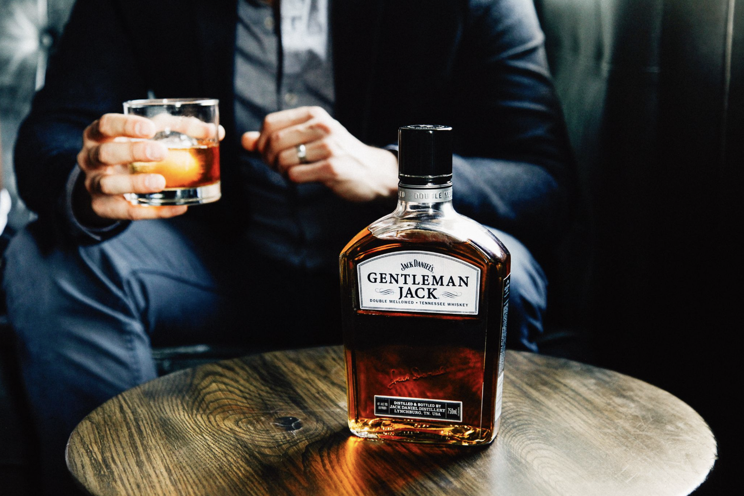 A person in a dark suit with a ring on their finger sitting at a wooden table, holding a glass of whiskey in their right hand, with a bottle of Jack Daniel's Gentleman Jack whiskey on the table.