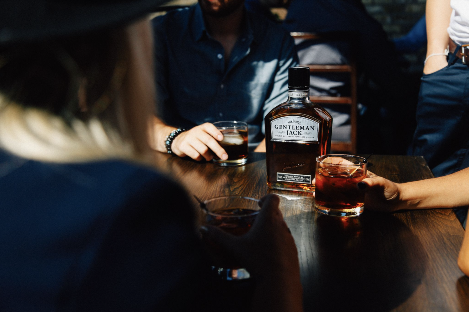 People sitting around a table with glasses of dark liquor and a bottle of Jack Daniel's Gentleman Jack whiskey.