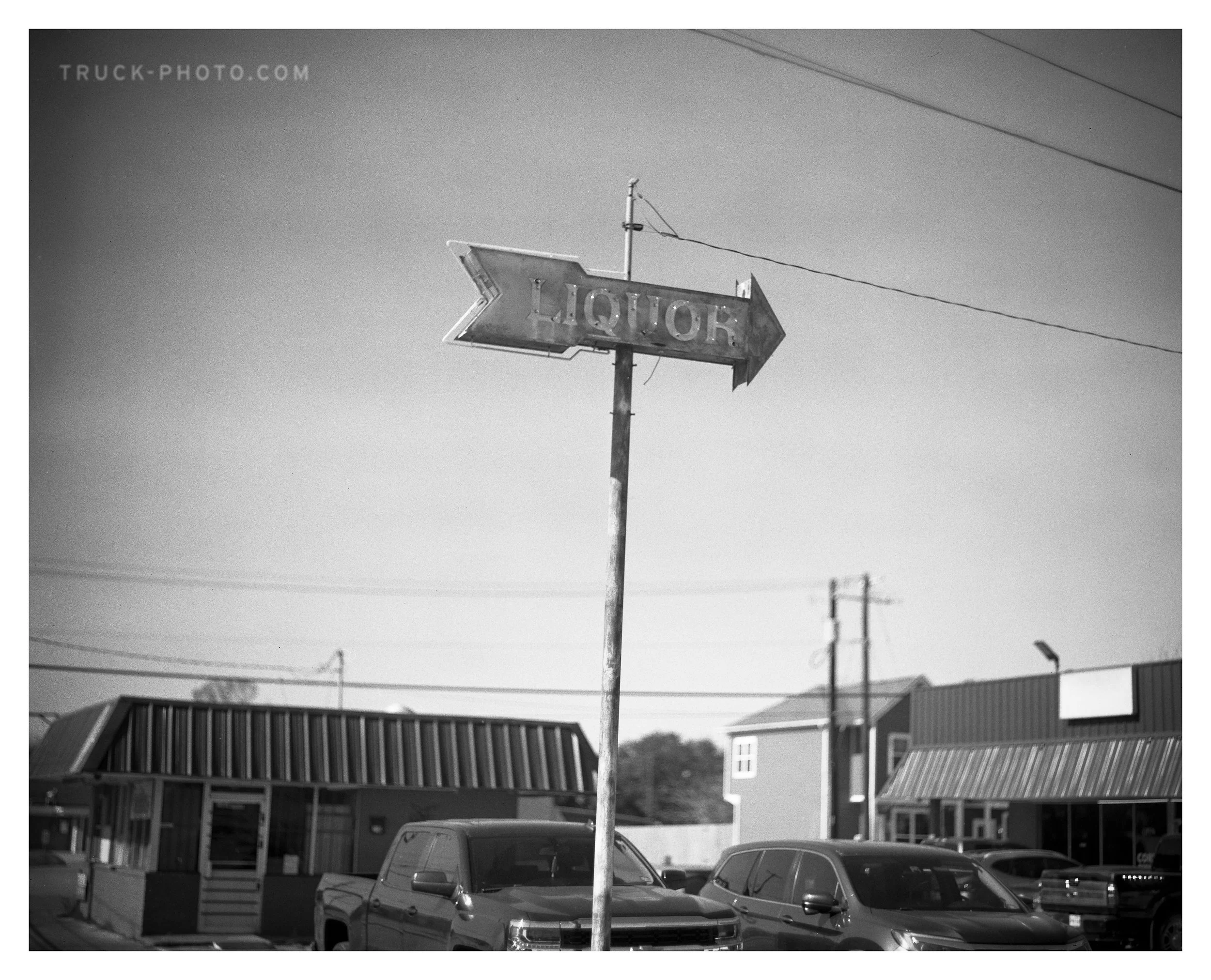 A worn vintage sign with the word 'LIQUOR' and an arrow pointing to the right, standing in a parking lot with modern cars and commercial buildings in the background.