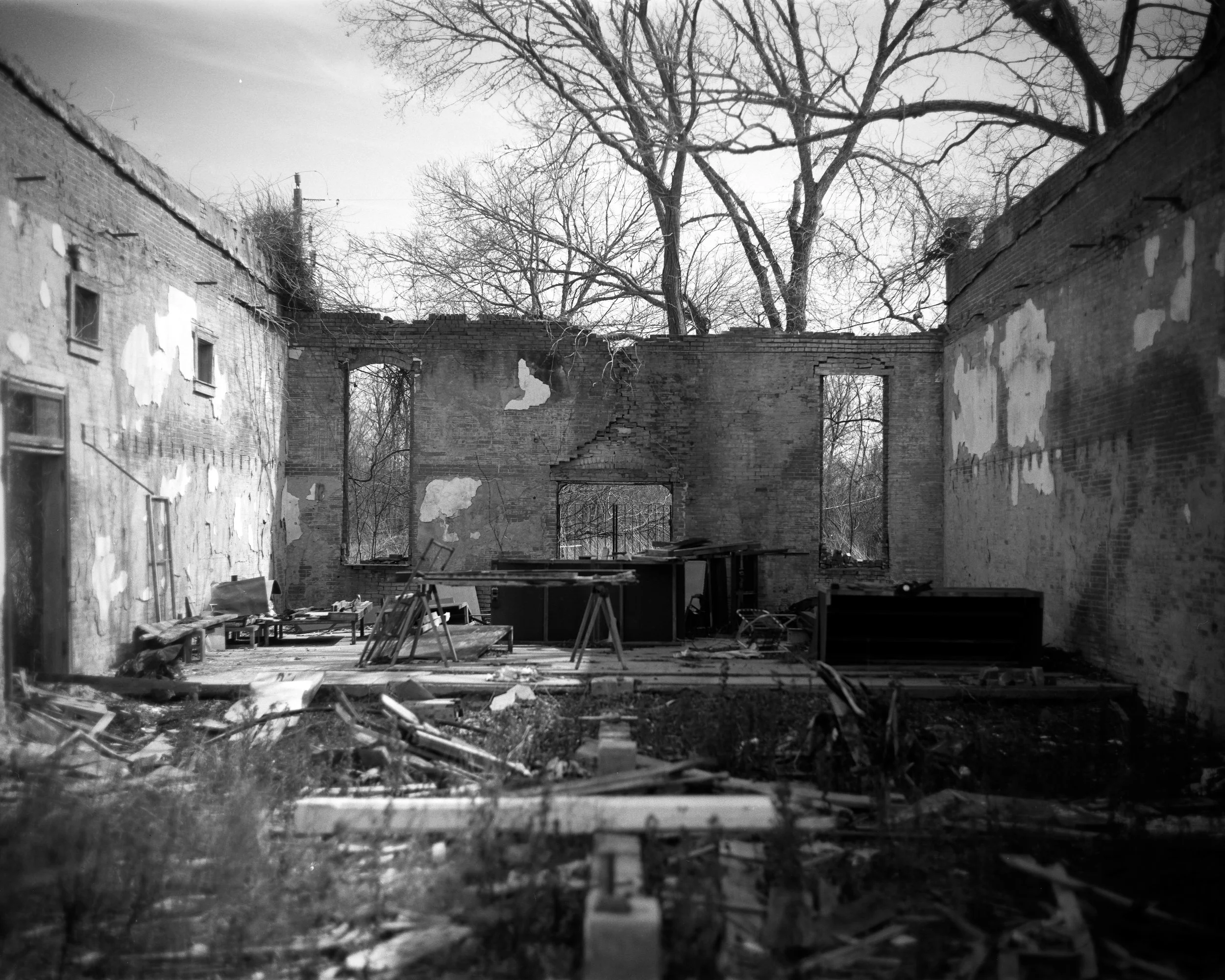 Black and white photo of a ruined brick building with missing walls and scattered debris inside, leafless trees visible through the empty windows and openings.