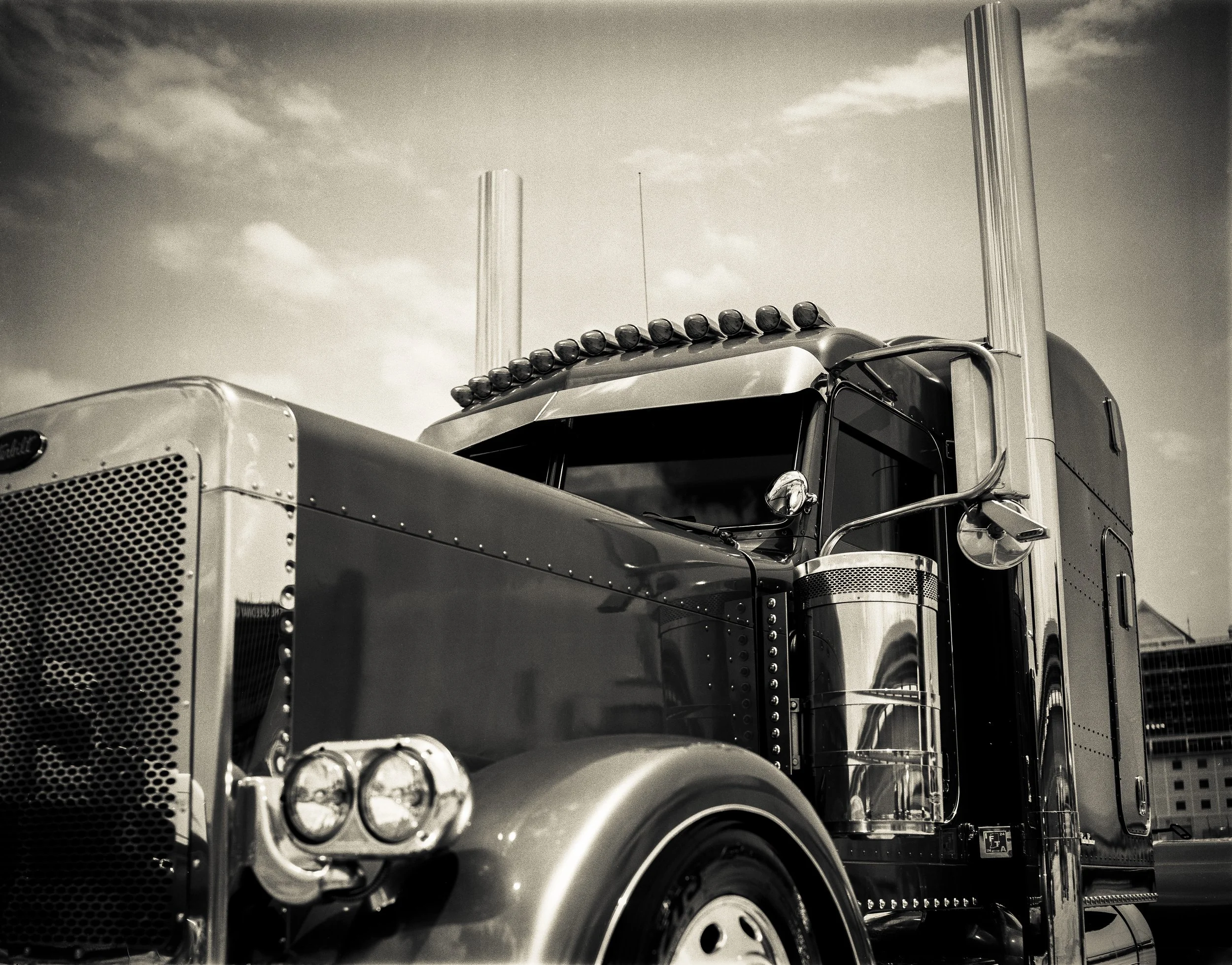 A black and white photograph of a large semi-truck with a prominent grille, rounded headlights, and tall exhaust pipes, viewed from the front-left side under a partly cloudy sky.