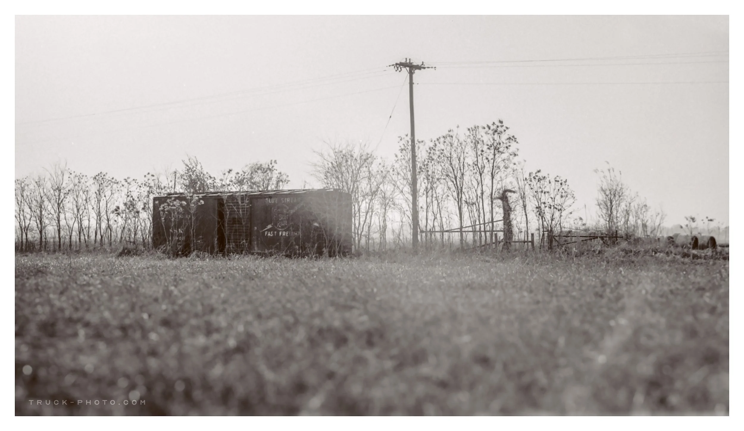 Black and white photo of an abandoned boxcar in a rural field with sparse trees, a utility pole, and a wooden fence.