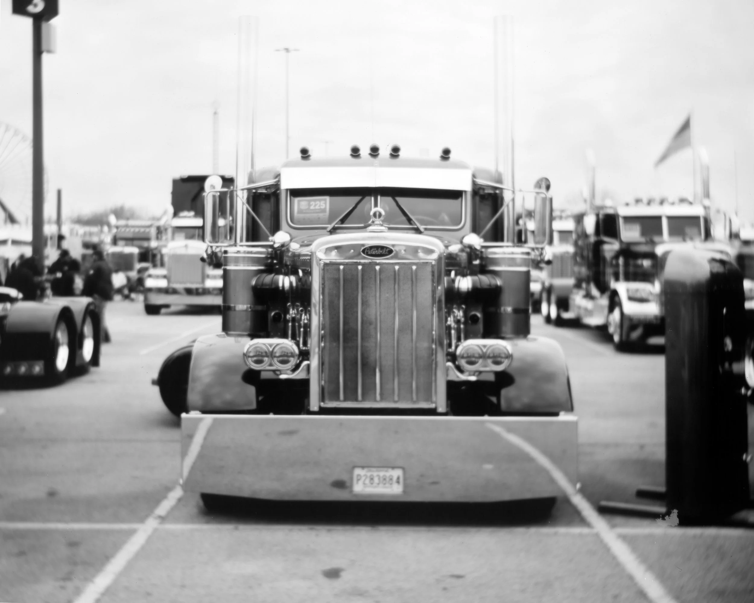 Black and white photograph of a vintage semi-truck parked in a lot with other vehicles, American flags in the background, and a striped barrier in the foreground.
