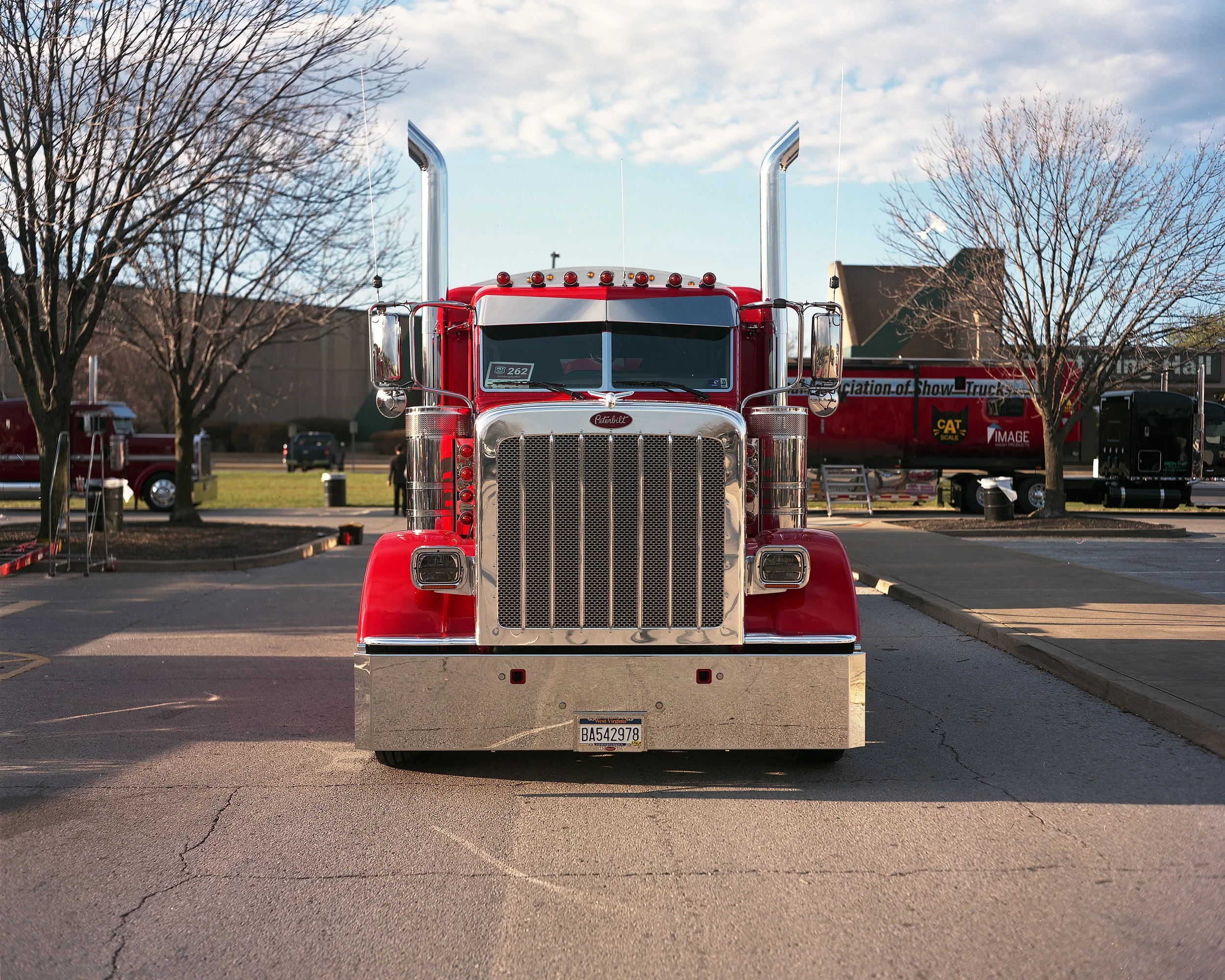 Front view of a large red Peterbilt parked in a lot with leafless trees in the background and a partly cloudy sky.