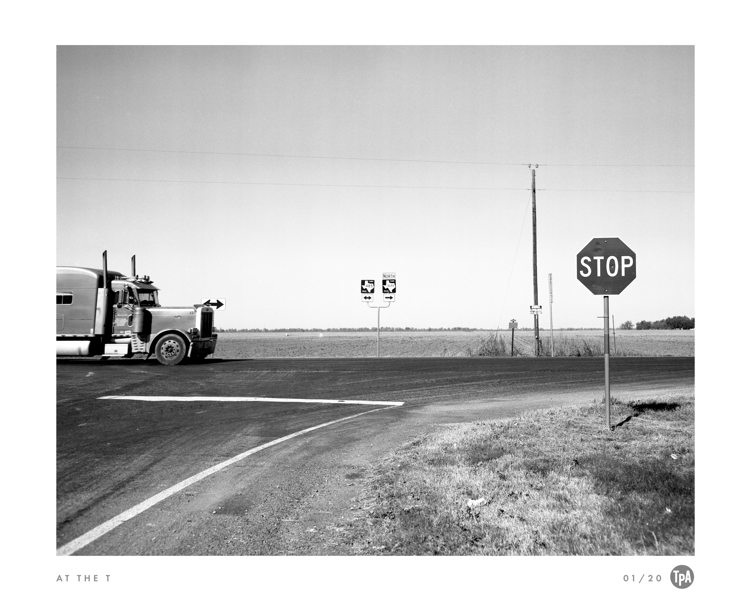 Black and white photograph of a rural road intersection with a truck, stop sign, utility pole, and highway signs indicating northbound routes.