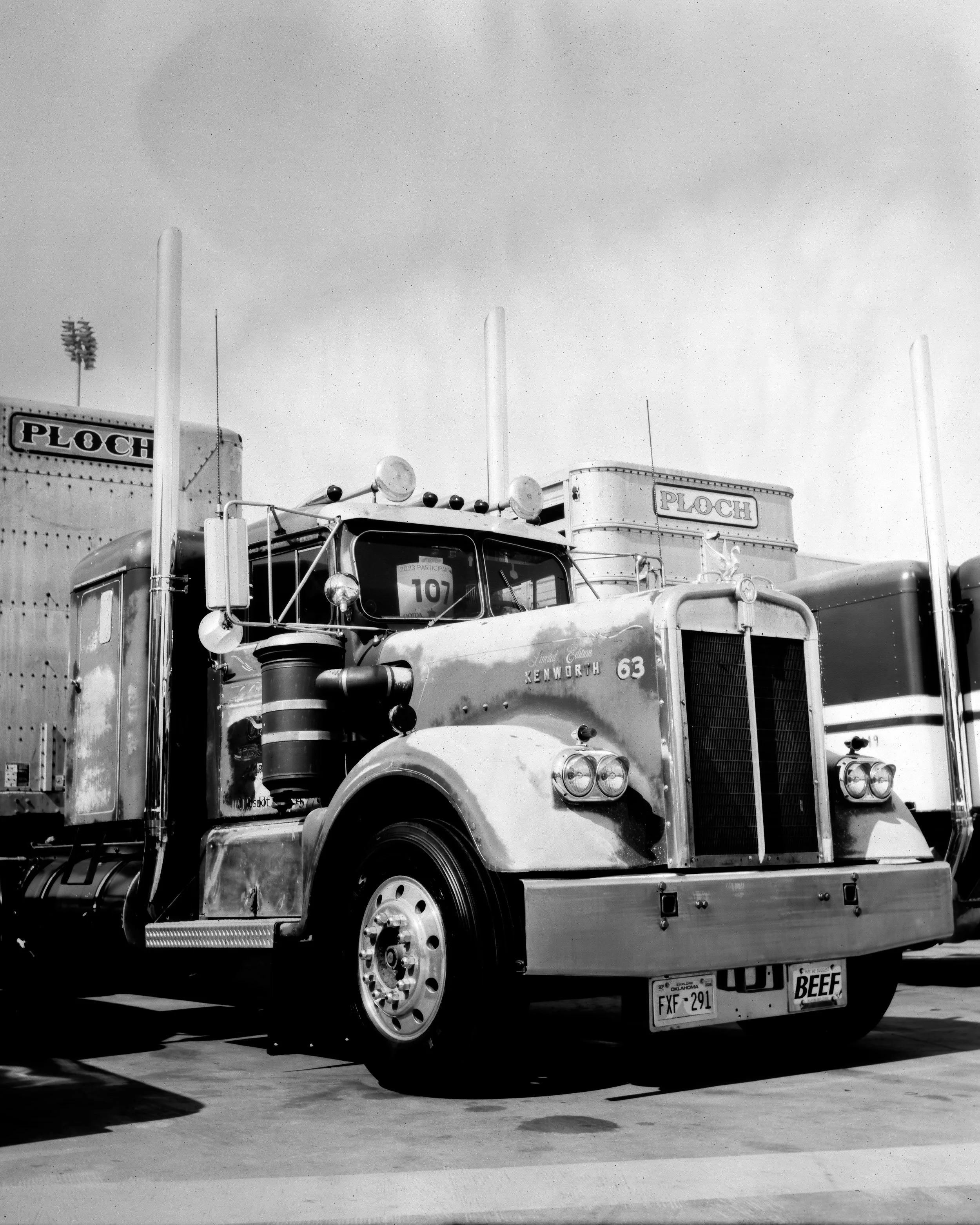 A black and white photo of a vintage Kenworth semi-truck with a weathered appearance, parked outdoors with trailers in the background. The truck has round headlights, dual air horns, and a prominent front grille.