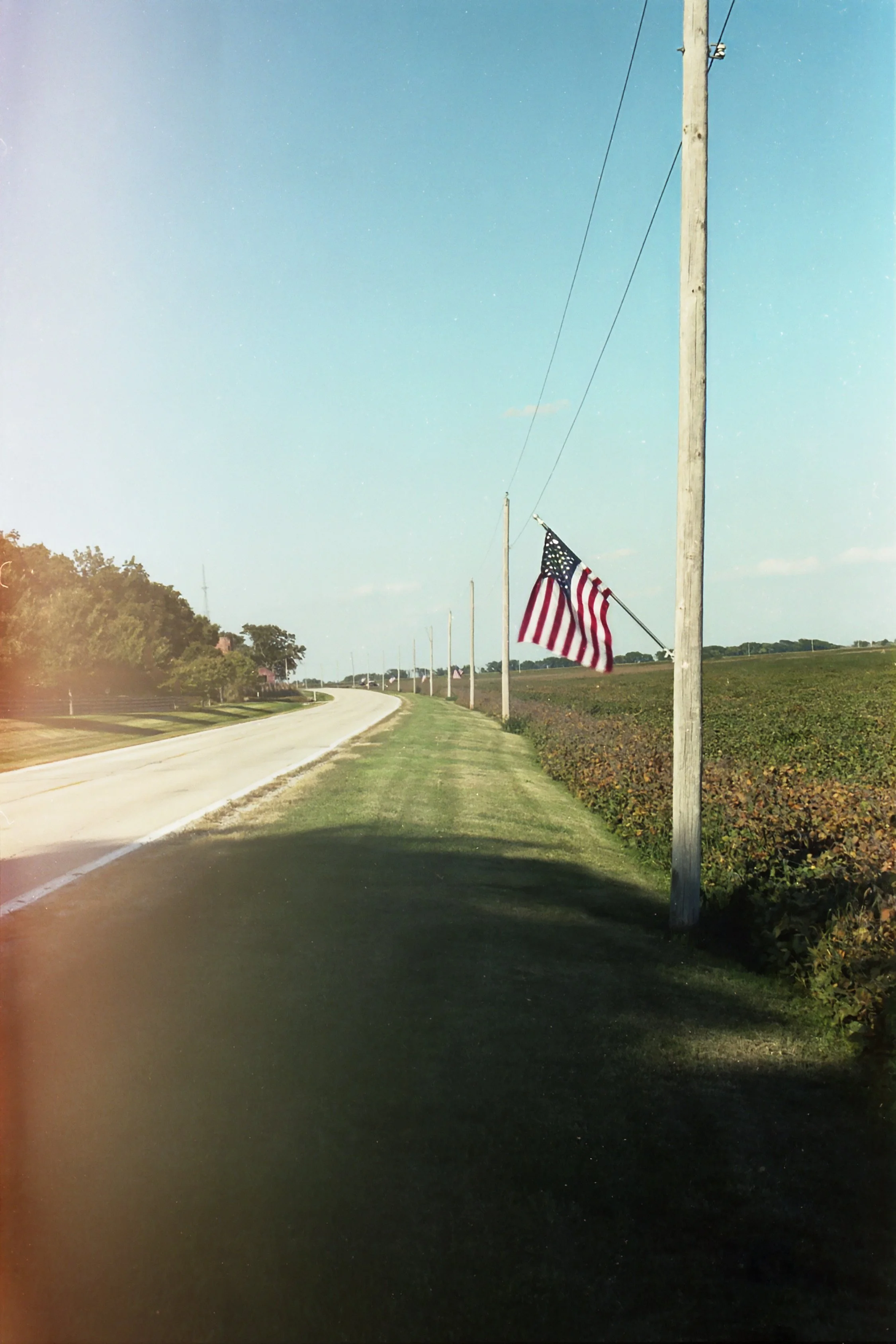 A rural road with an American flag on a pole on the right side, green fields, trees, and power lines in the background, under a clear blue sky.