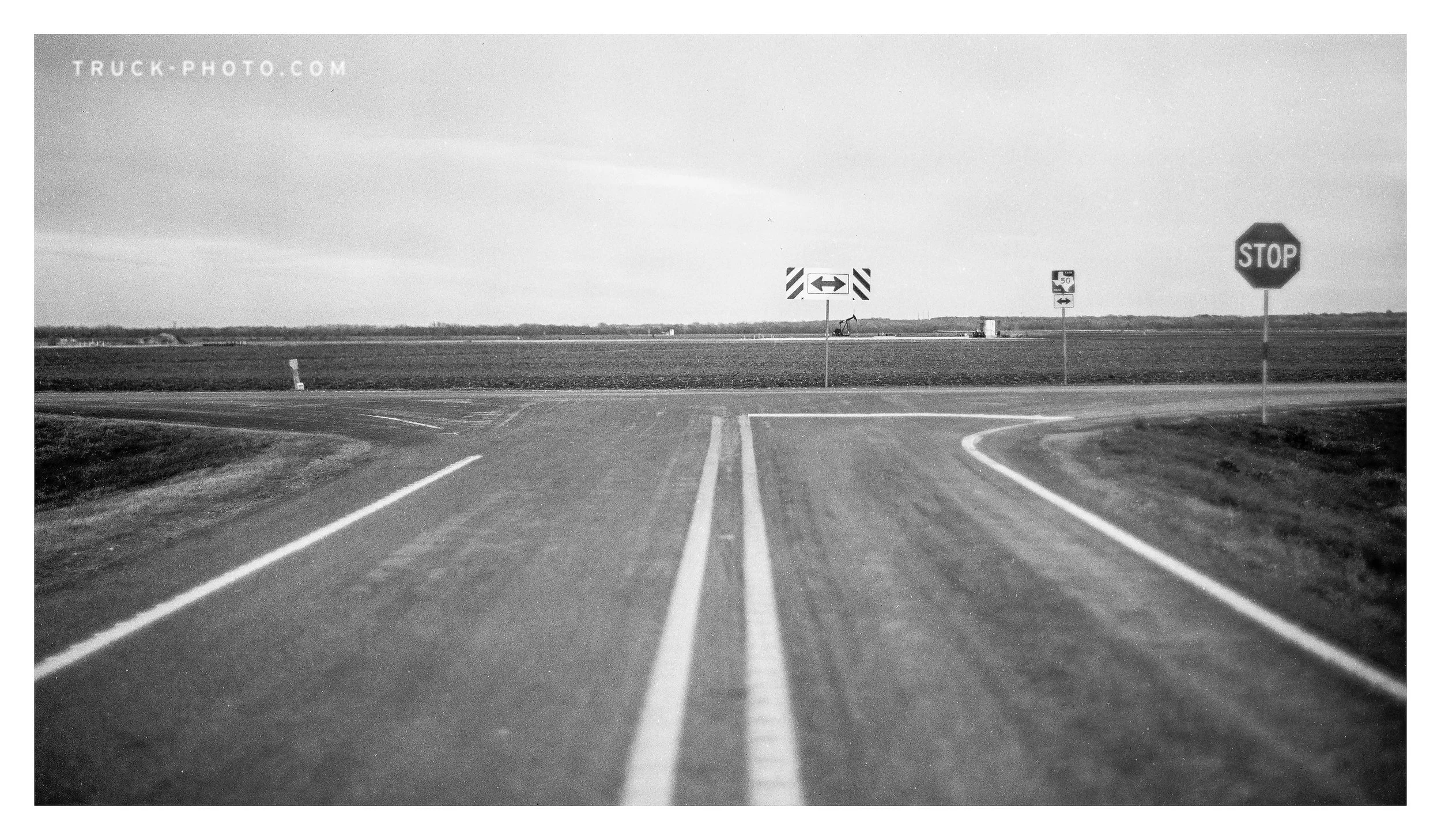 Black and white photo of a rural intersection with a stop sign on the right, road signs for left and right turns ahead, and an open field in the background.