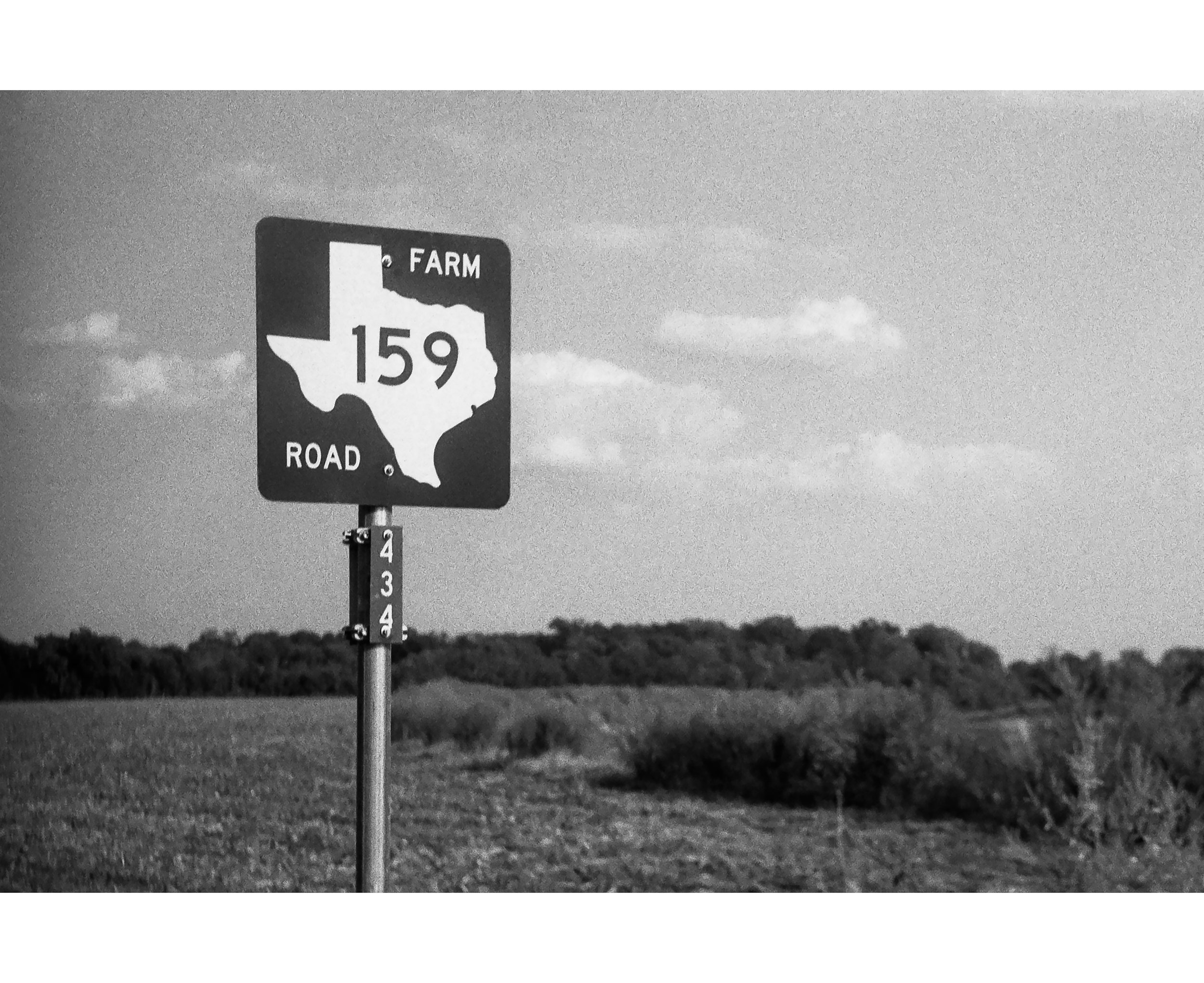 Black and white image of a road sign indicating Texas State Highway 159, with a map of Texas and the number 159, against a rural landscape with grass and trees in the background.