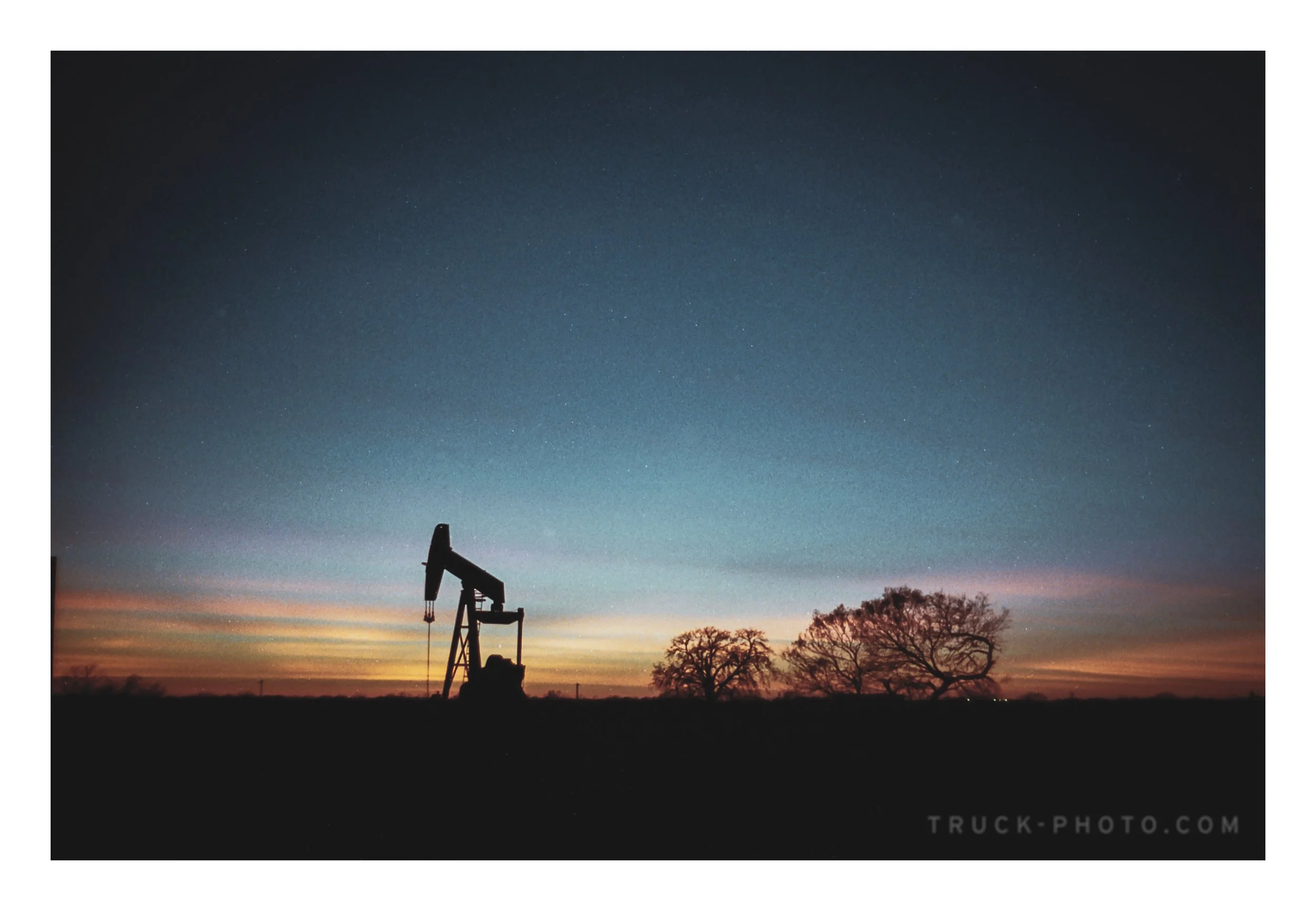 A silhouette of an oil pumpjack in a rural landscape at dusk, with a colorful sky and a few trees in the background.