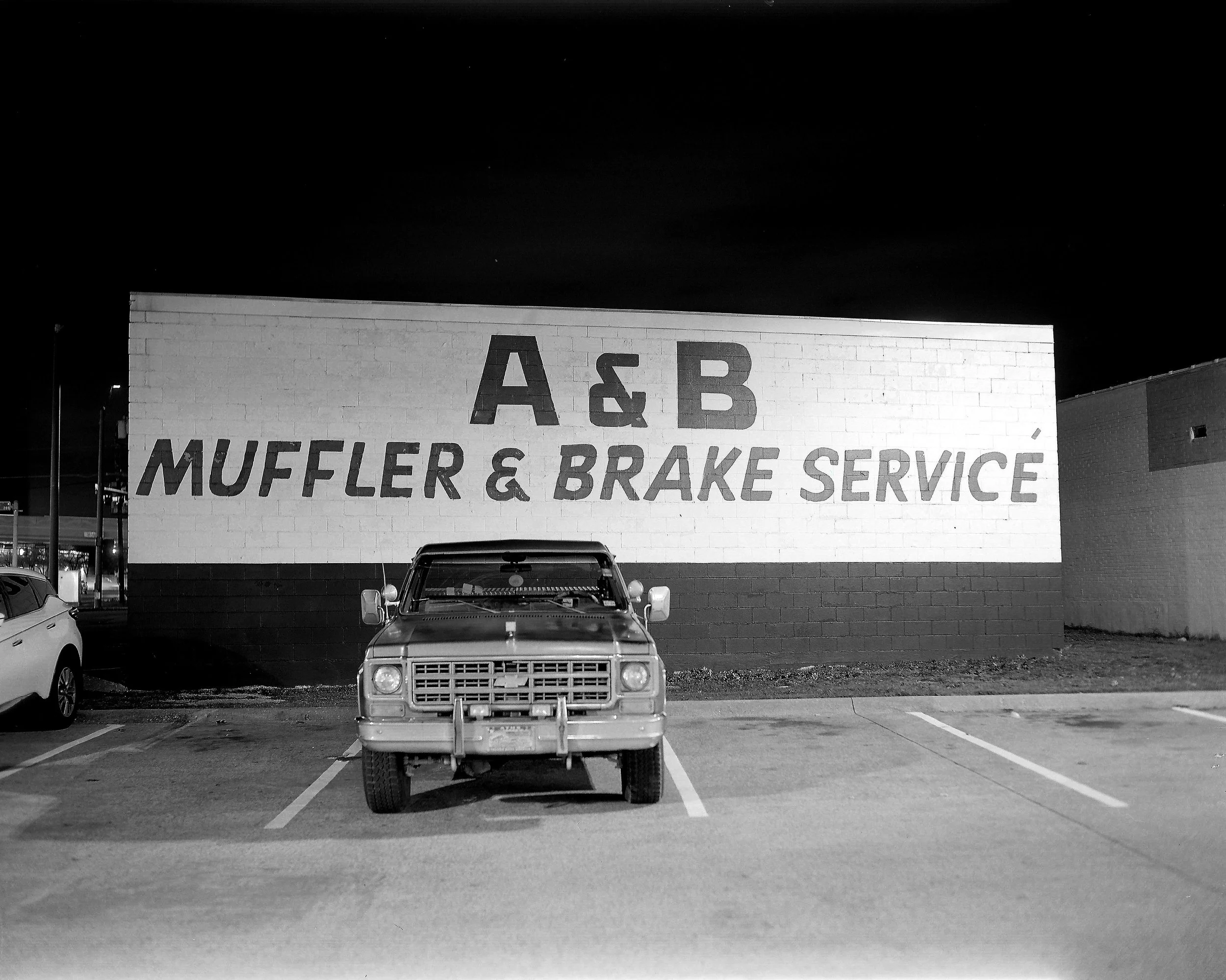 A parking lot at night with a vintage truck centered in front of a wall. The wall has the large text 'A & B MUFFLER & BRAKE SERVICE' painted on it.