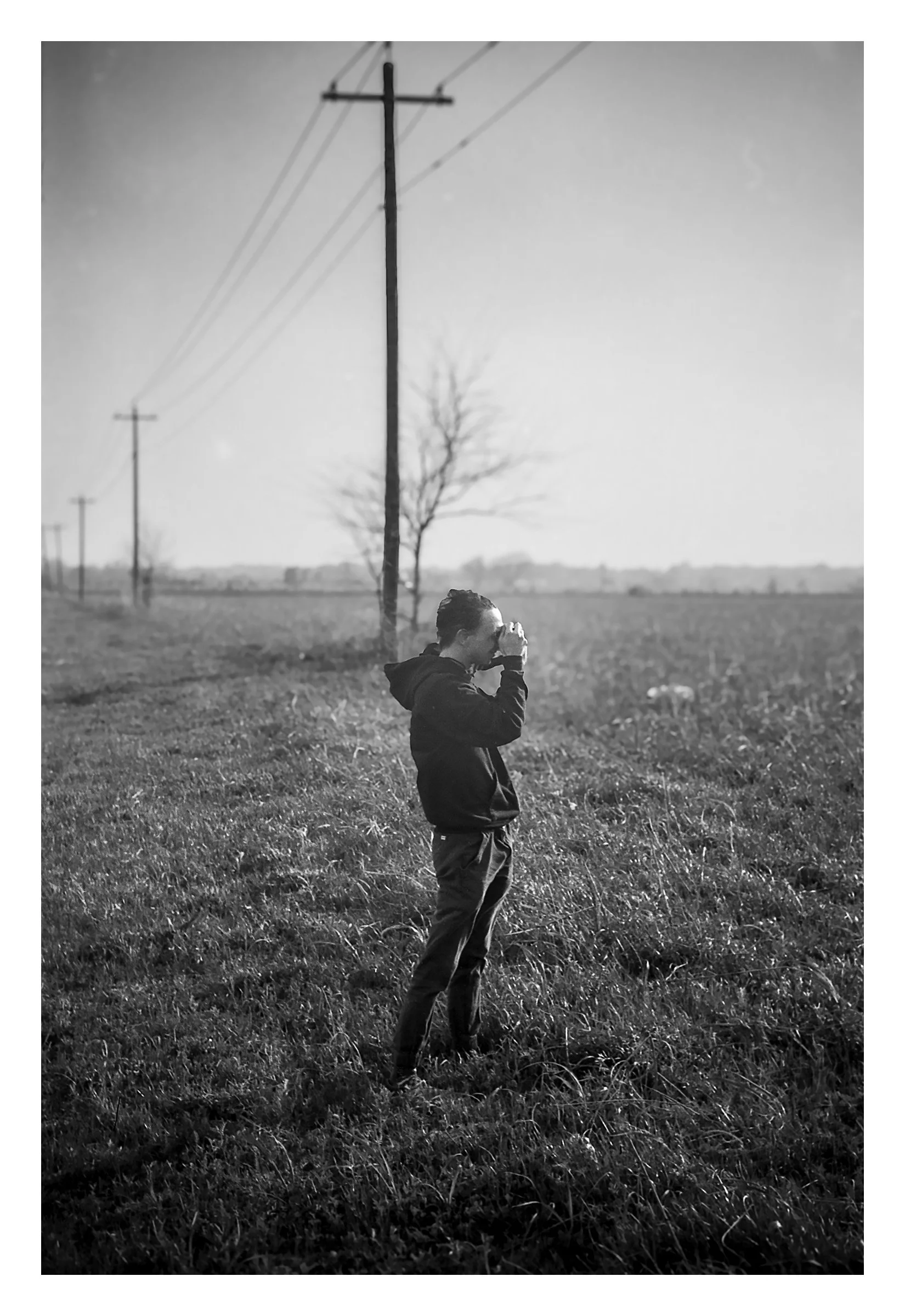 A person standing in a grassy field near power lines, taking a photograph, with a tree and open landscape in the background.