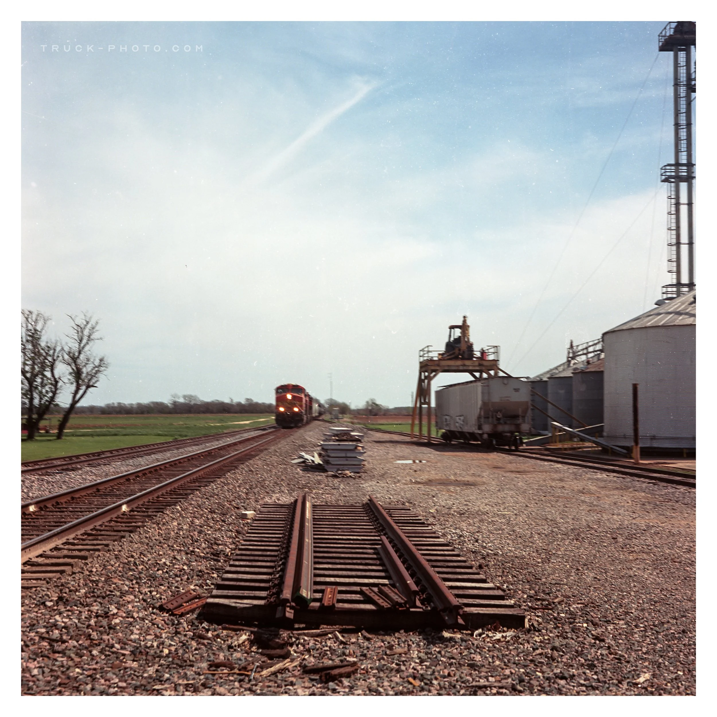 A train approaching on railway tracks near a rural industrial site with silos and equipment, under a partly cloudy sky.