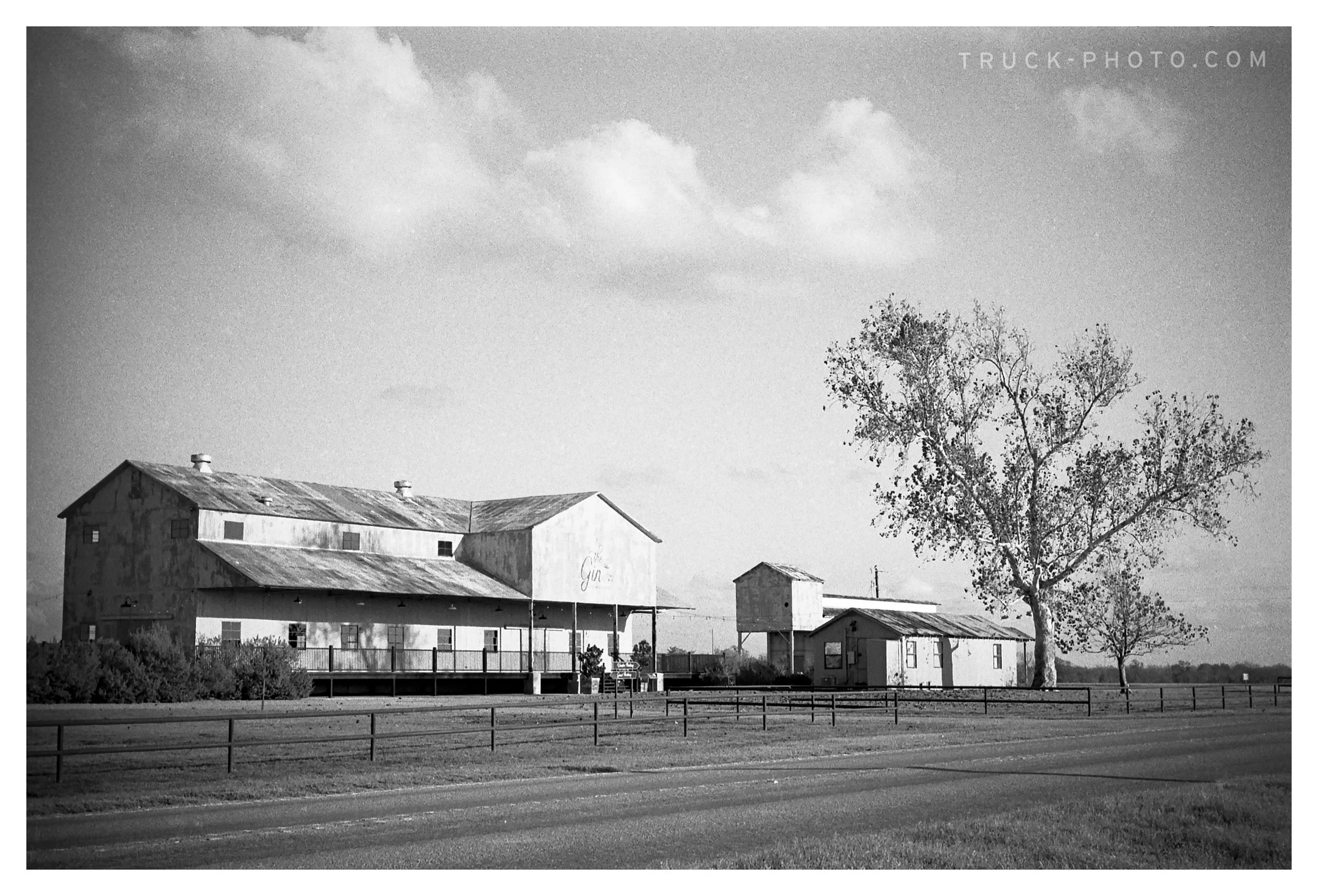 Black and white photo of an old farmstead with a large barn, smaller sheds, and a tree in the foreground. The farm buildings have a weathered appearance with metal roofs, and the landscape is open with a field and clouds in the sky.