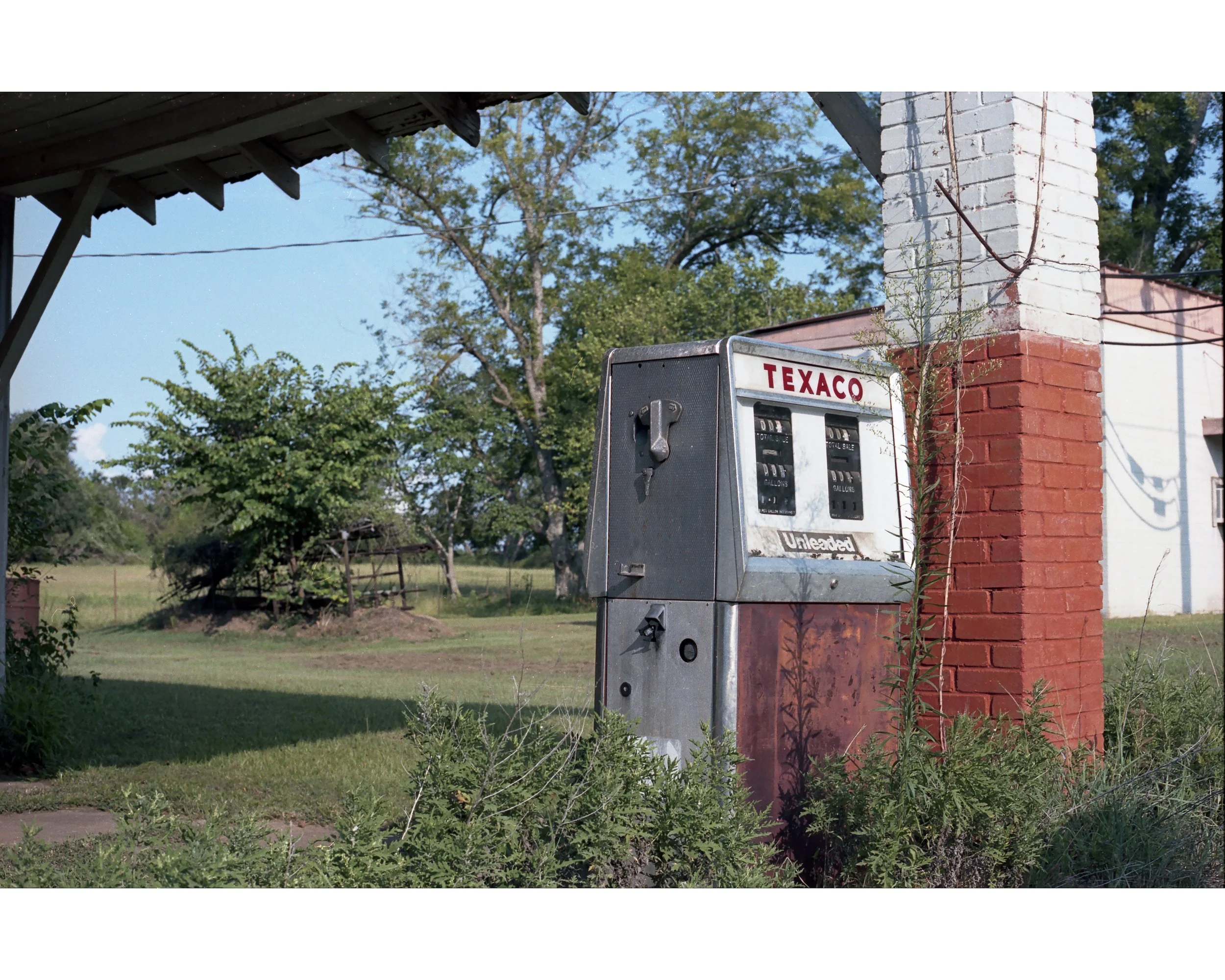 An old Texaco gasoline pump near a brick and white painted building, surrounded by grass, weeds, and trees in a rural area.
