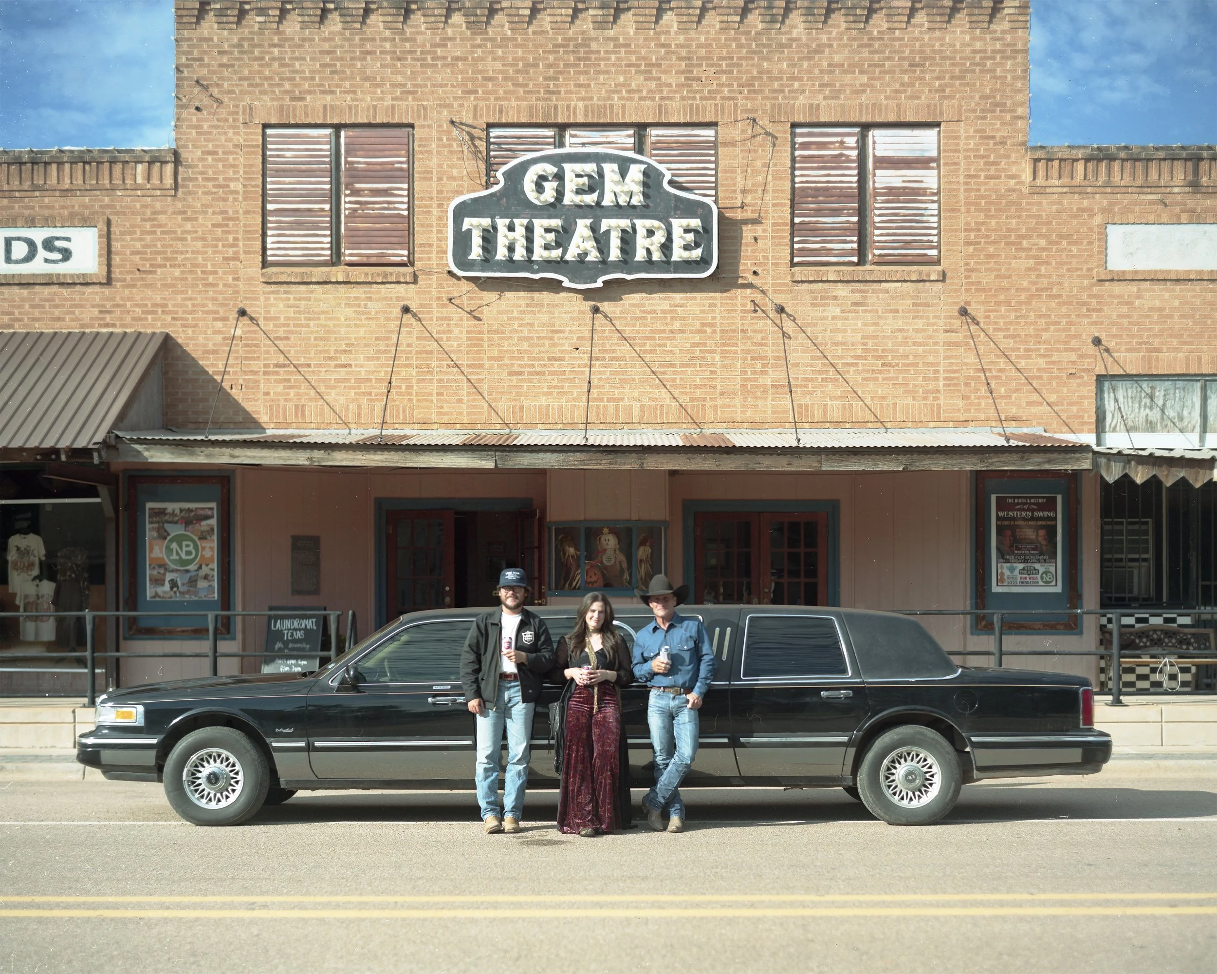 Three people standing in front of a black limousine parked on a street in front of the Gem Theatre building, with a brick facade and vintage posters.