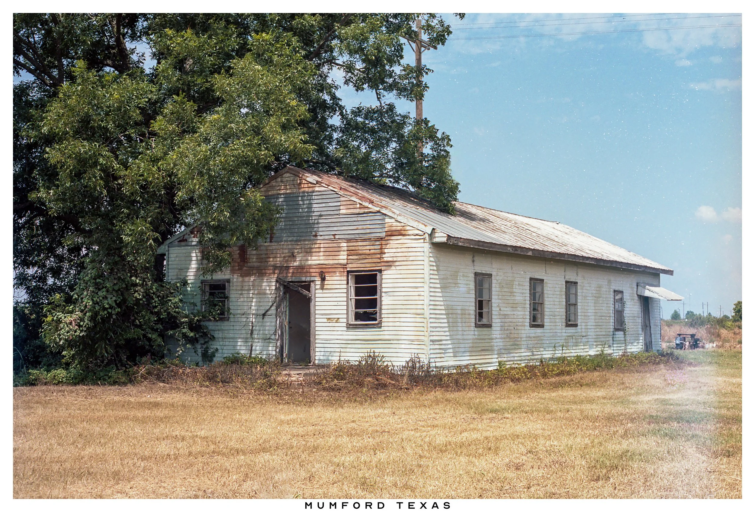 An old, weathered white house with peeling paint, rusted roof, and broken windows, surrounded by a dry grassy field and a large tree in Mumford, Texas.