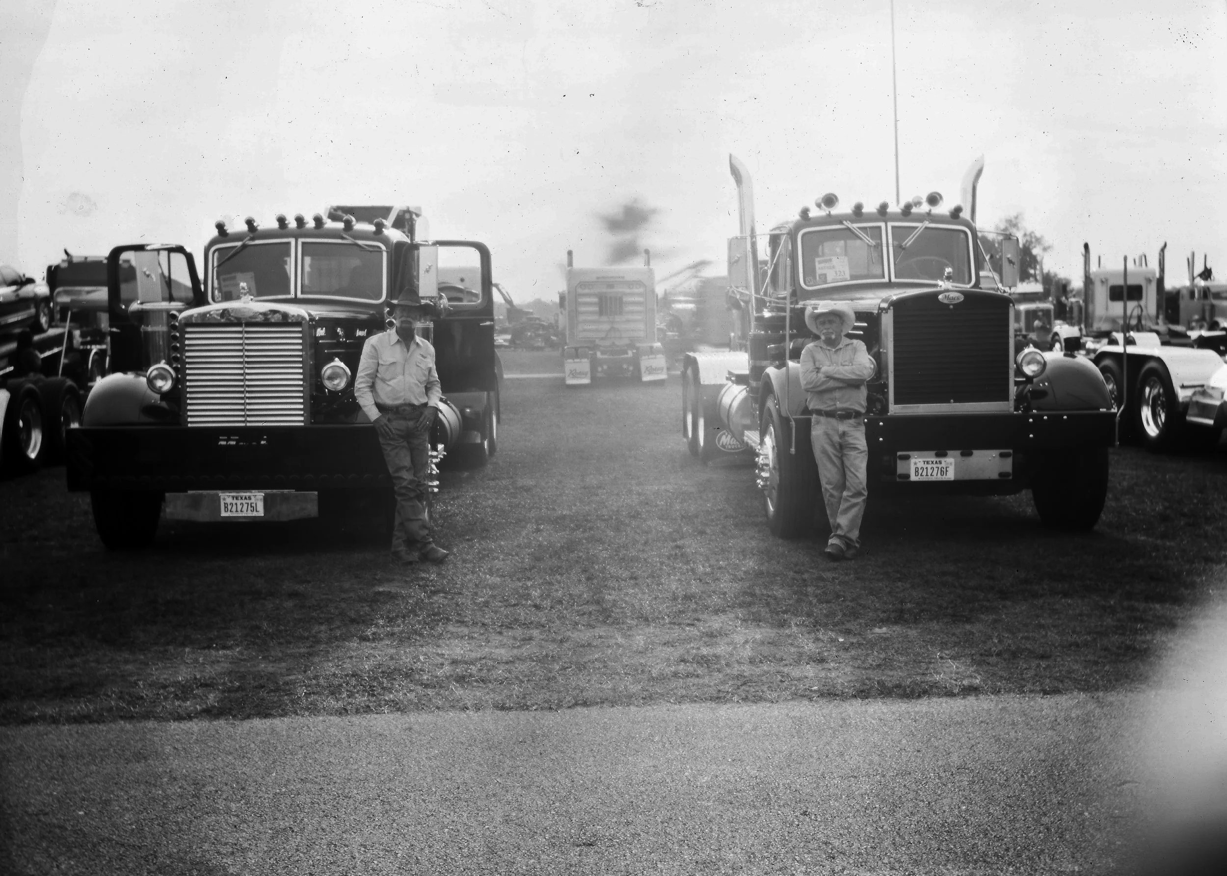 Two men standing beside large semi trucks at a truck show or lot, black and white photograph.