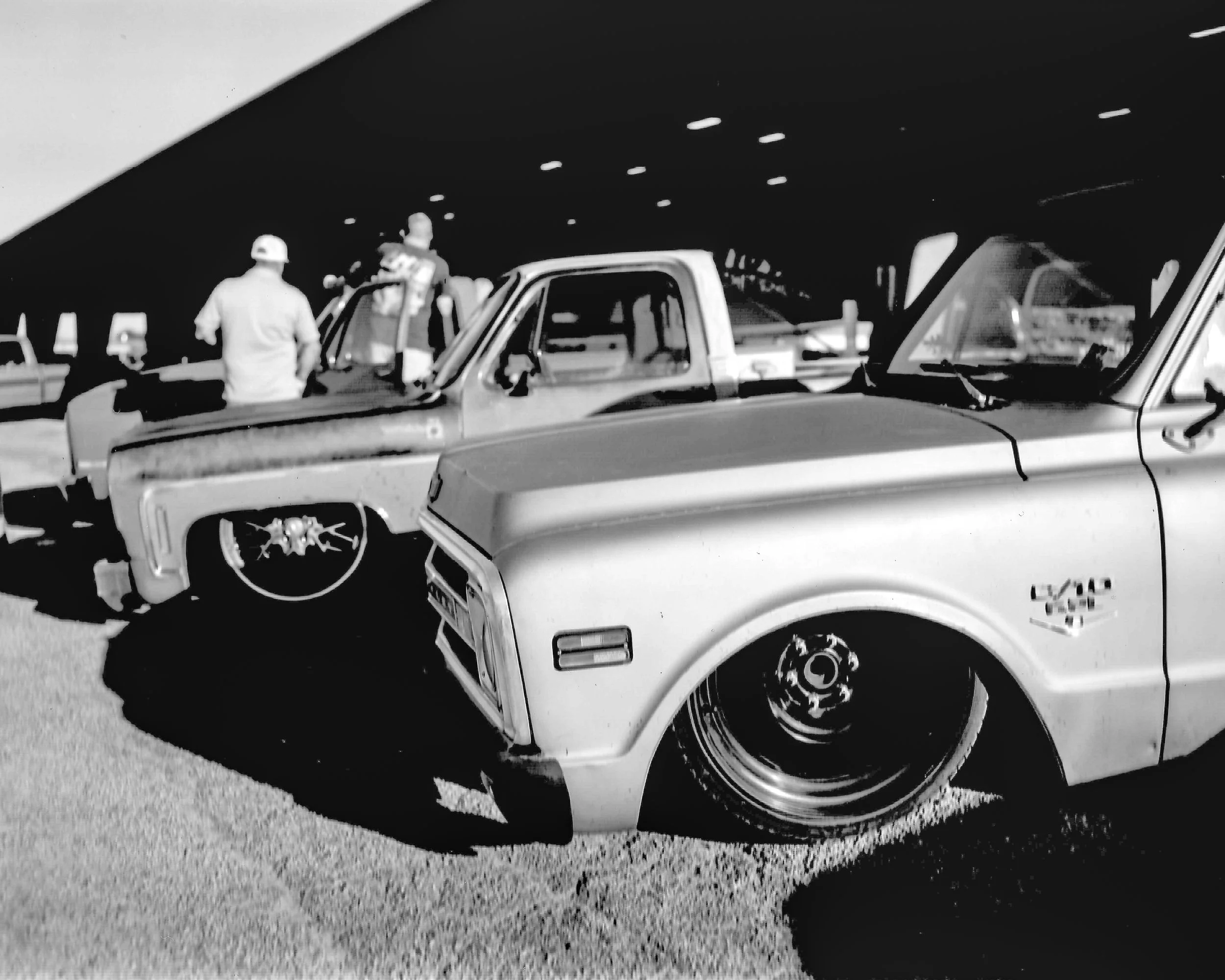 Black and white photo of two pickup trucks parked under a bridge, with three men nearby, one with a cap, and the trucks have custom rims and low-profile tires.
