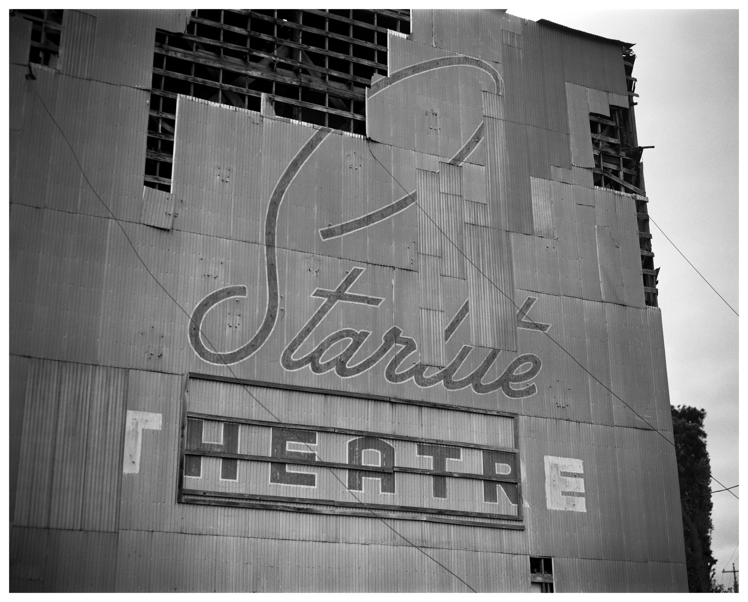 A black-and-white photograph of a worn, corrugated metal building with a vintage sign that reads 'Starlite' in script and 'THEATRE' in block letters beneath it. The building shows signs of disrepair with missing panels and exposed structural elements