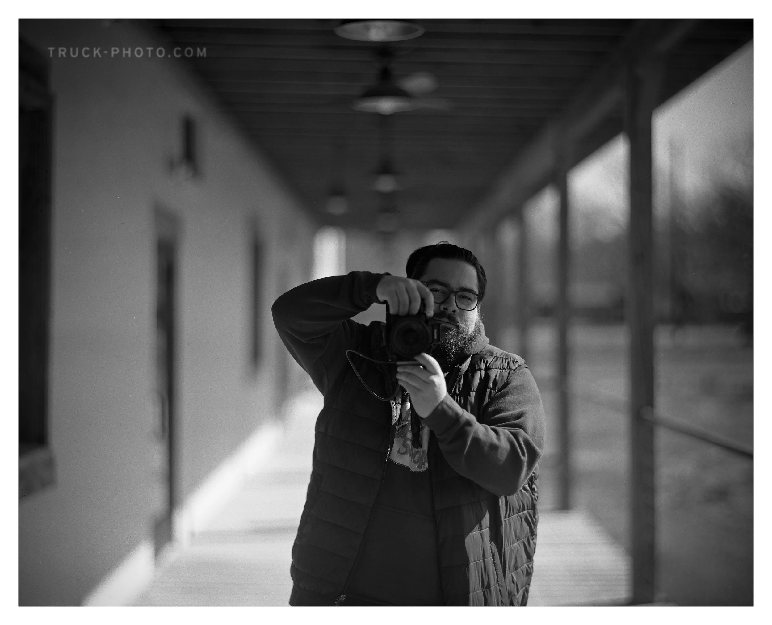 A man takes a selfie in a mirror on a porch, holding a camera, wearing glasses and a padded vest, with blurred background of the porch and outdoors.