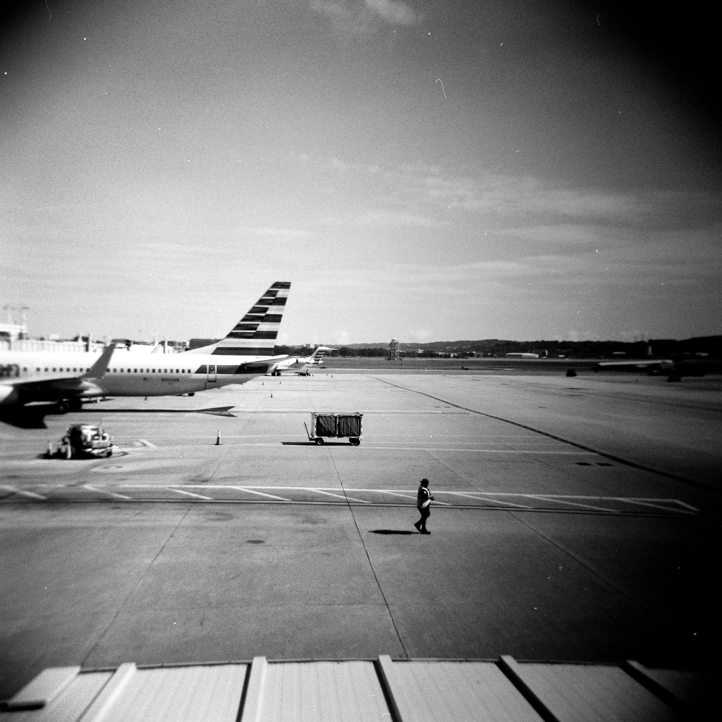 Black and white photo of an airport tarmac with airplanes parked, a ground support vehicle, and a person walking.