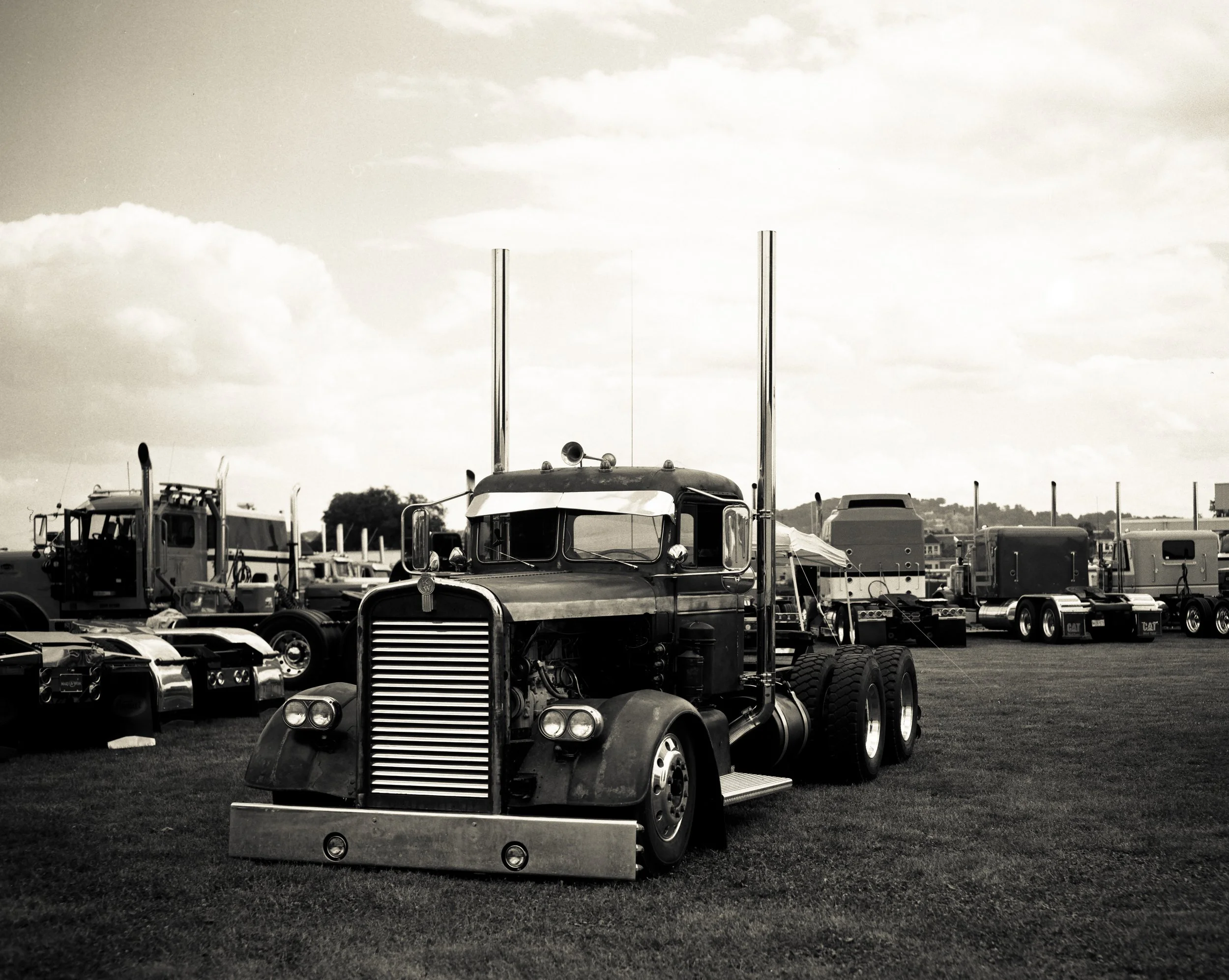 A black and white photo of a classic semi-truck parked on grass with other trucks in the background.