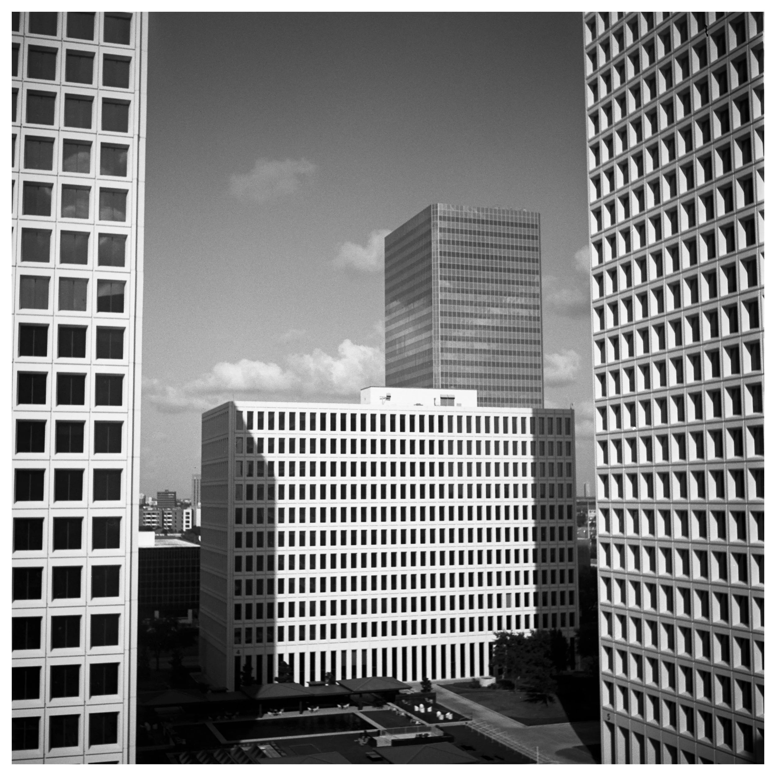 Black and white photo of modern cityscape with tall office buildings, including two facades with grid-like windows, and a taller building in the background under a partly cloudy sky.